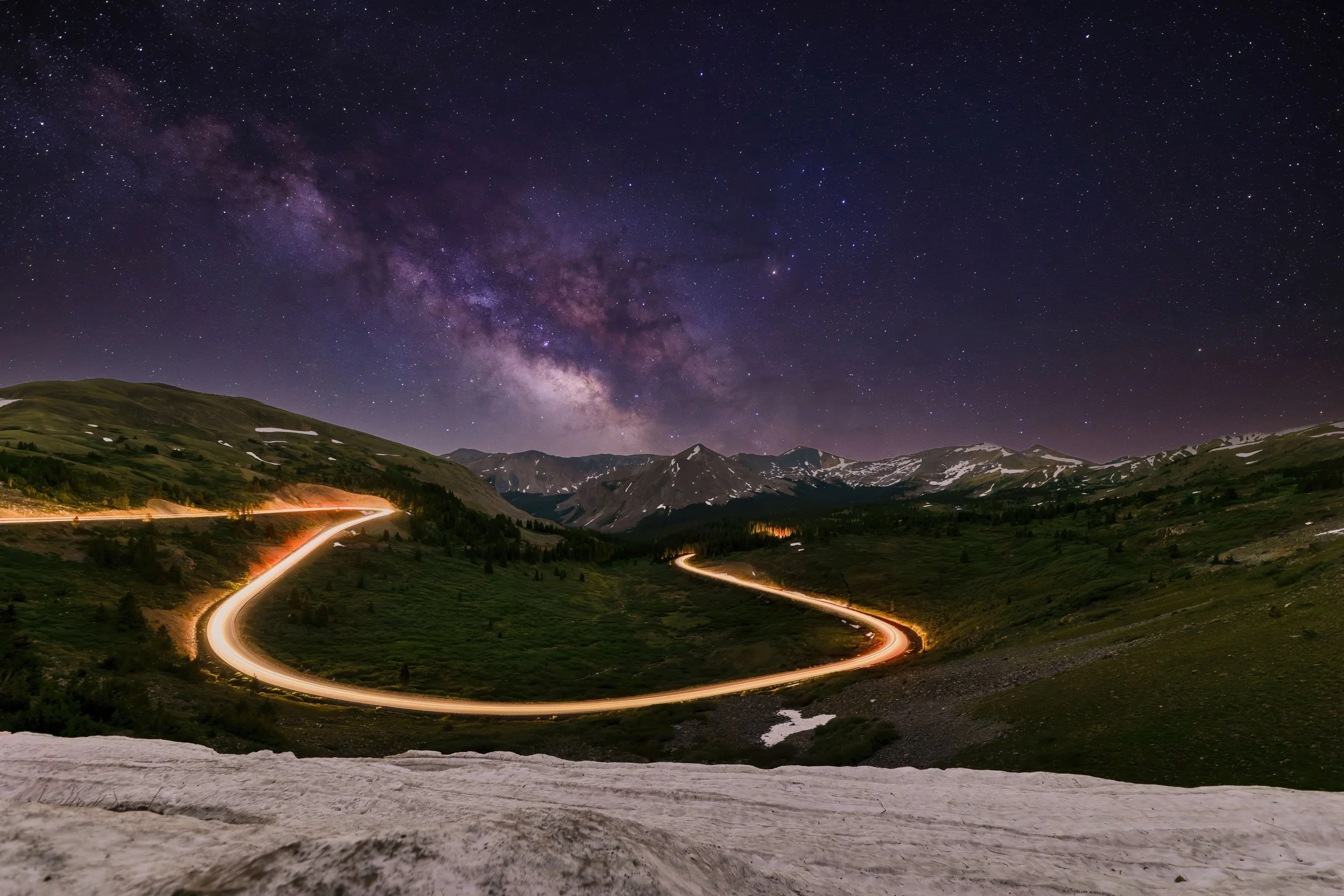 Nighttime landscape of a mountainous area with a winding road illuminated by car lights, under a star-filled sky and the Milky Way galaxy.