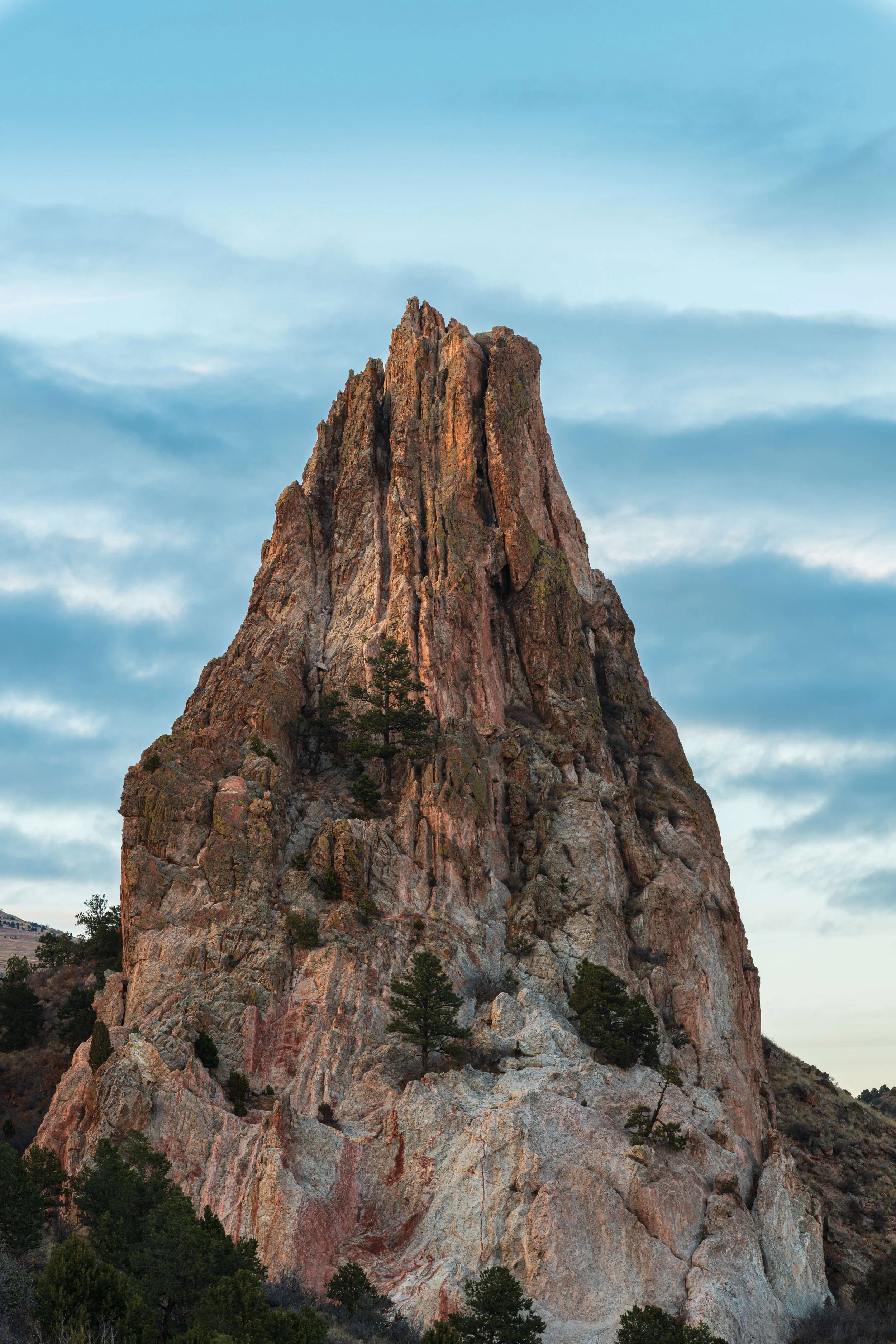 A large, rugged rock formation rising into a partly cloudy sky, with sparse trees growing at its base and on its slopes.