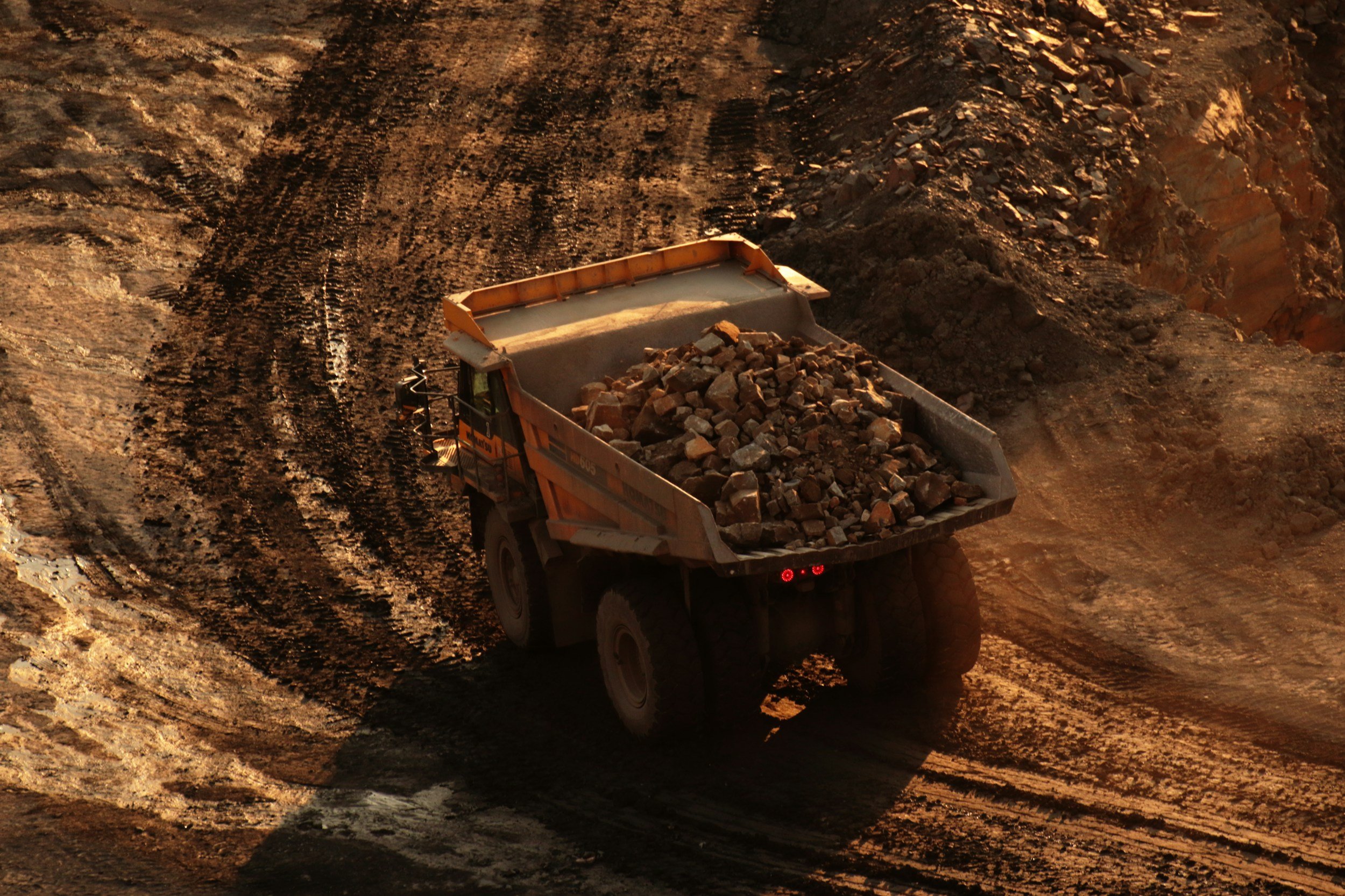 A large construction dump truck filled with rocks moving along a dirt road at a construction site, surrounded by rugged terrain.