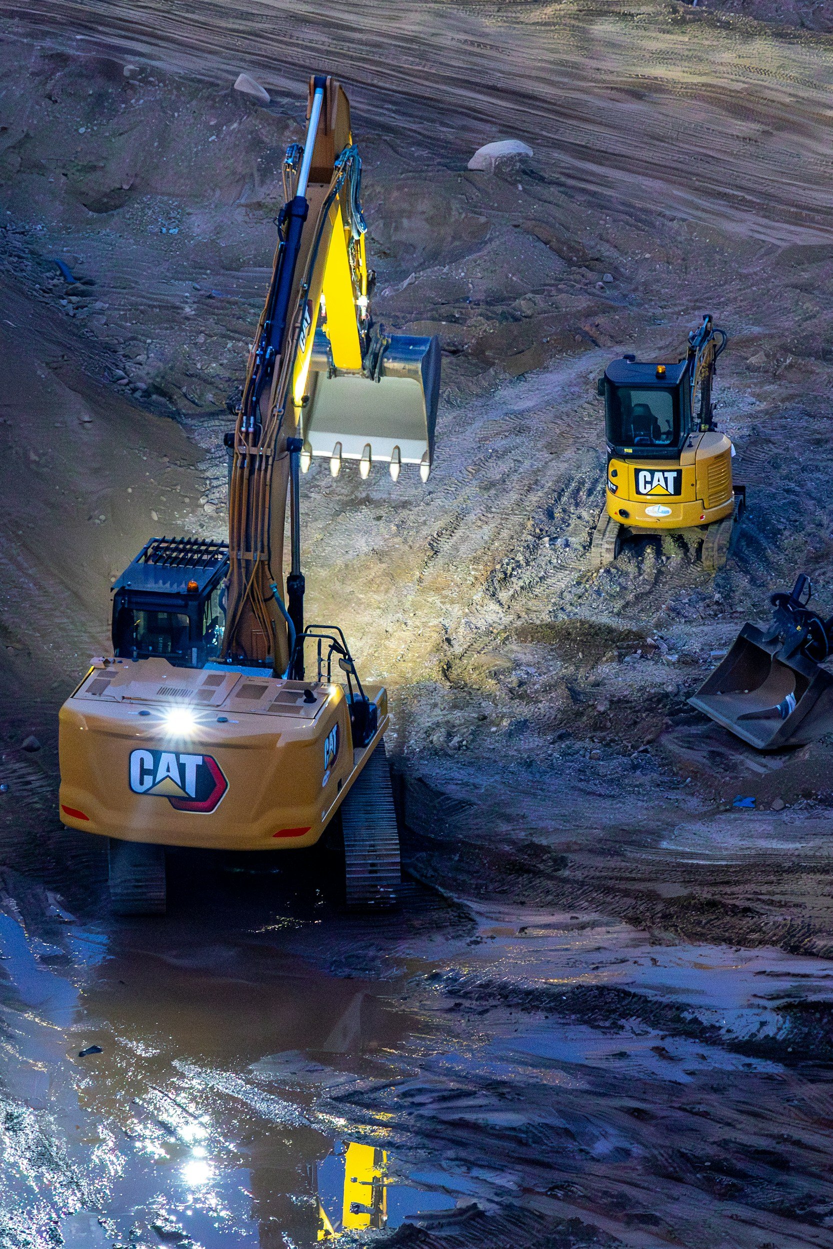 Two yellow Cat construction excavators working on a muddy construction site at night, illuminated by bright lights, with one excavator digging and the other parked nearby.