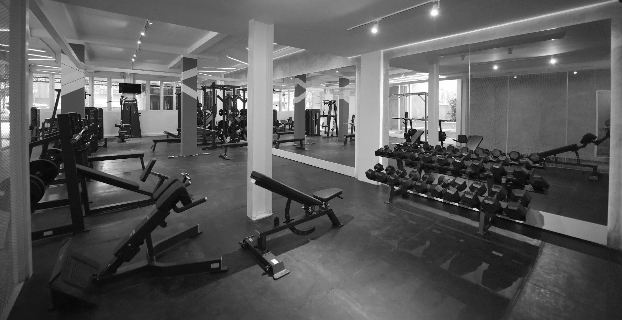 Interior of a modern gym with various workout equipment including dumbbells, weight benches, and cardio machines, illuminated by ceiling lights and large windows.