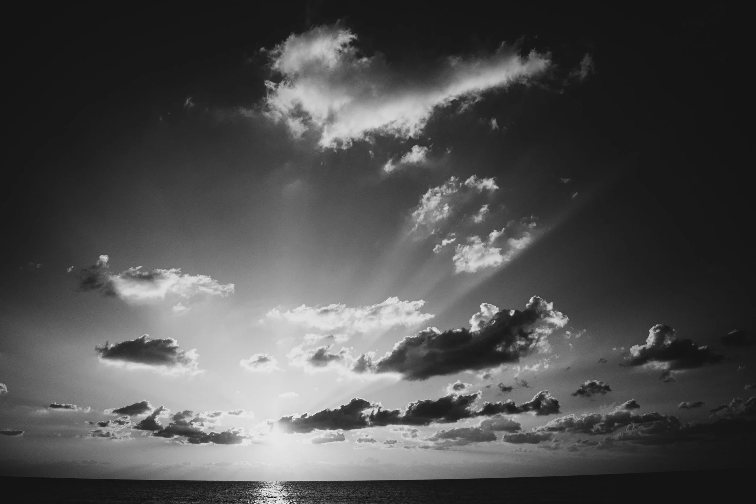 Black and white photo of a sunset over the ocean with clouds in the sky and sunlight breaking through.