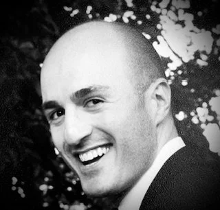 Black and white photo of a smiling man with a shaved head, wearing a suit, outdoors with trees in the background.