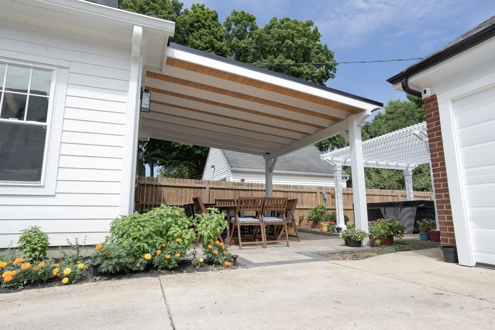 Backyard patio with white house siding, wooden fence, potted plants, and outdoor dining table with chairs under a covered patio.