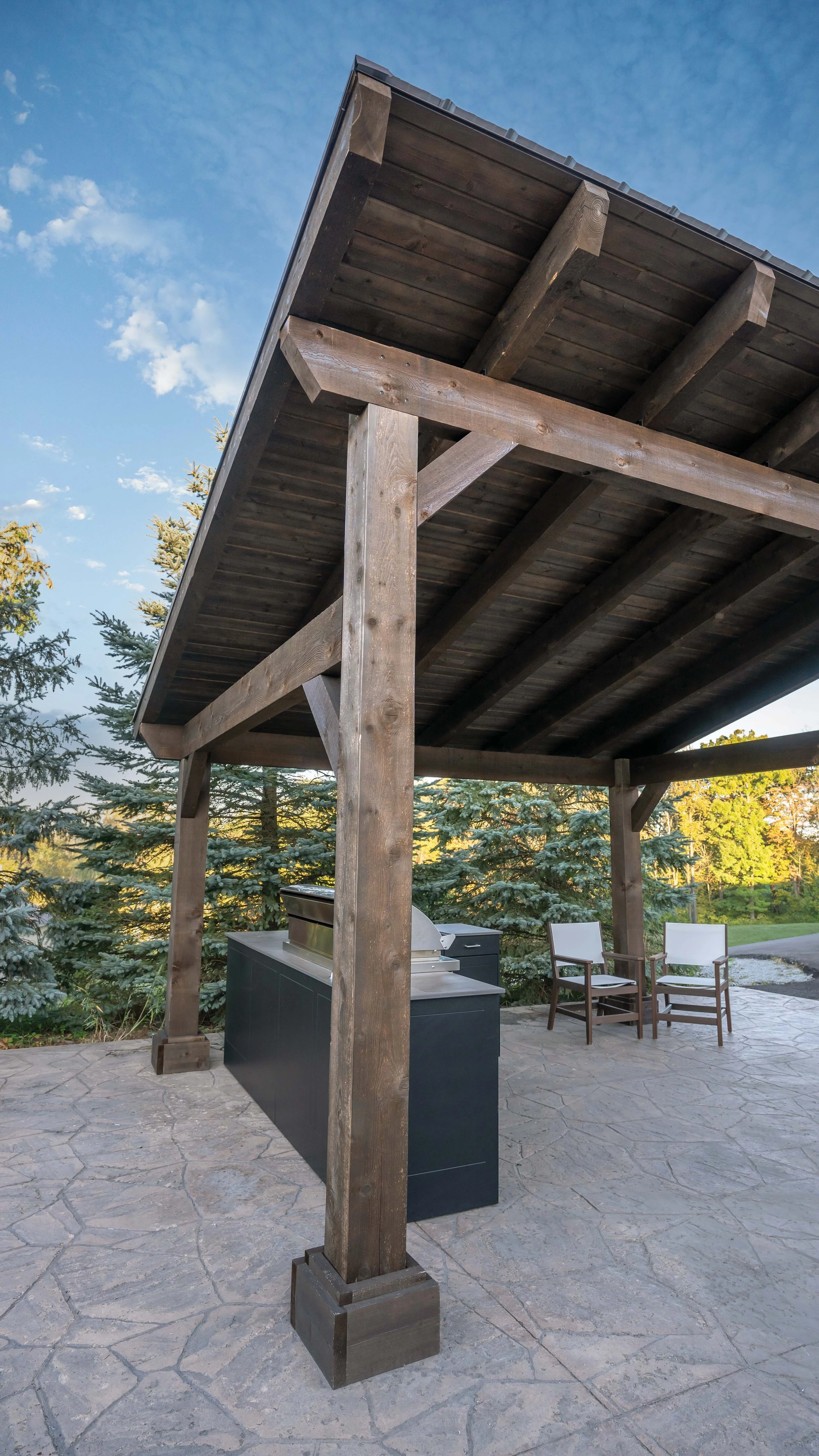 An outdoor covered patio with a built-in grill, two chairs, and a stone floor, surrounded by trees.