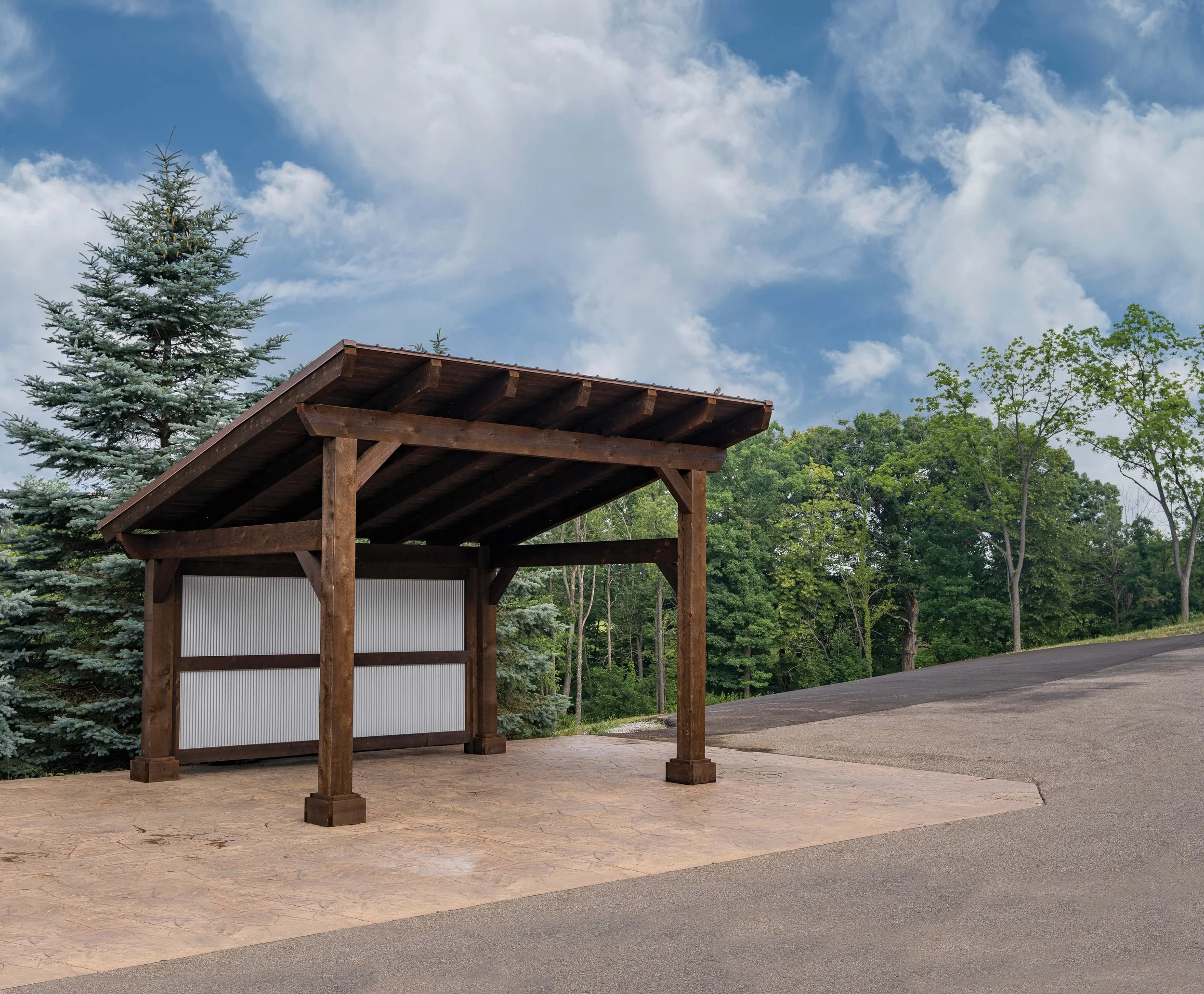 A wooden shelter with a slanted roof, situated on a paved area next to a paved road, with green trees and a partly cloudy sky in the background.