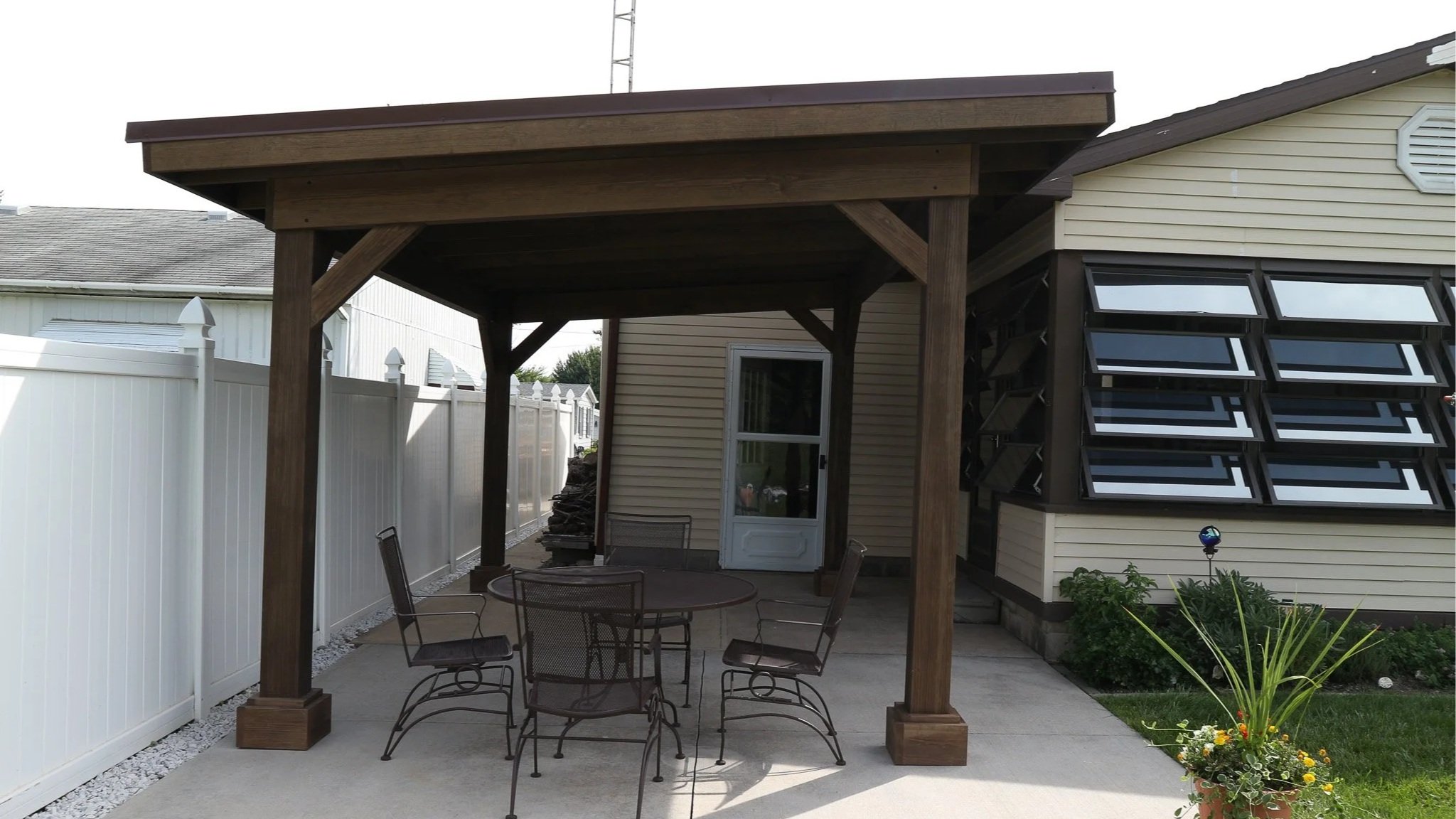 Backyard patio with a wooden covered pergola, outdoor dining table with five chairs, white privacy fence, potted flowers, and a house with multiple small windows.