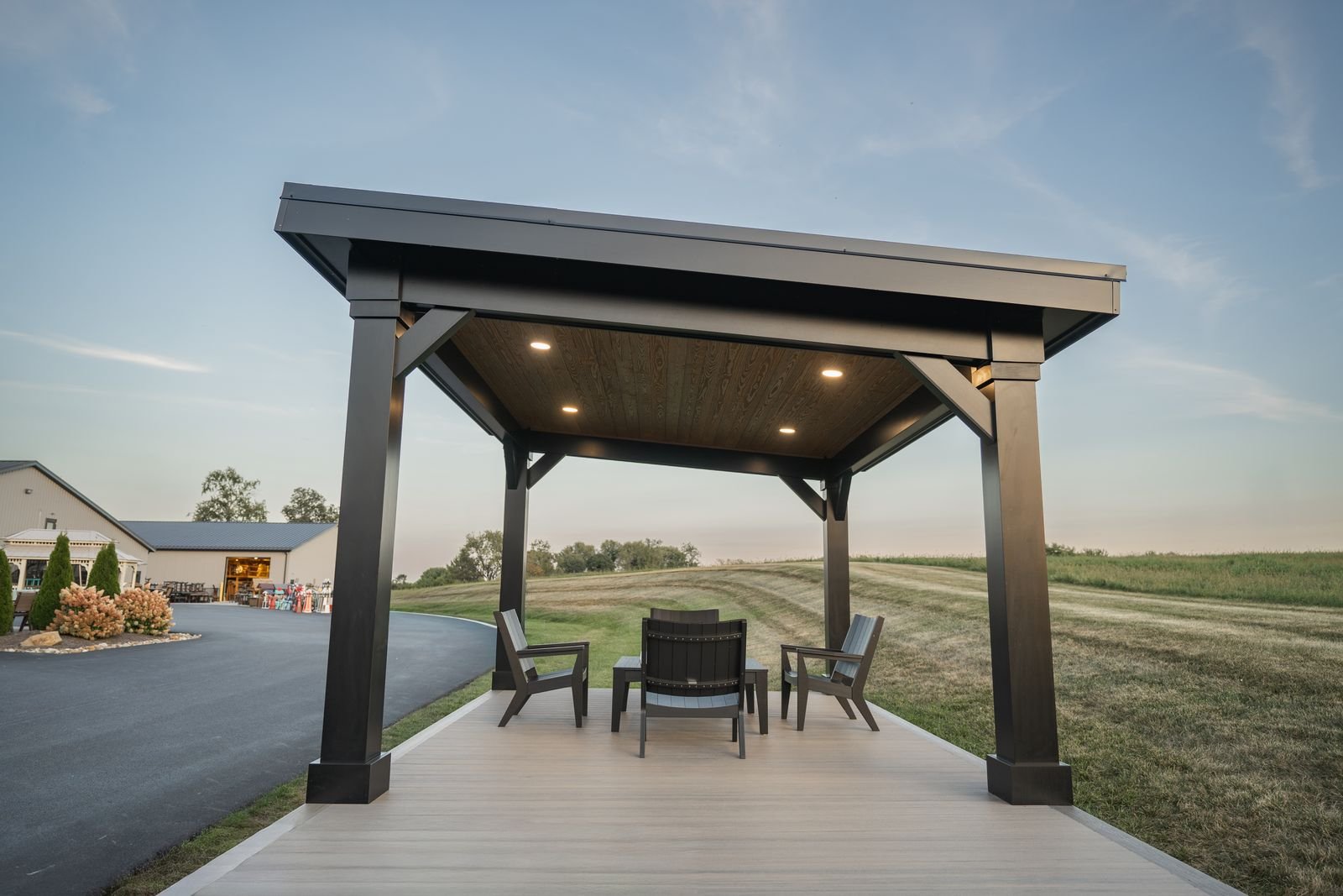 Empty outdoor pavilion with four black chairs and a small table on a light wooden deck, overlooking grassy fields and a building in the distance during early evening.