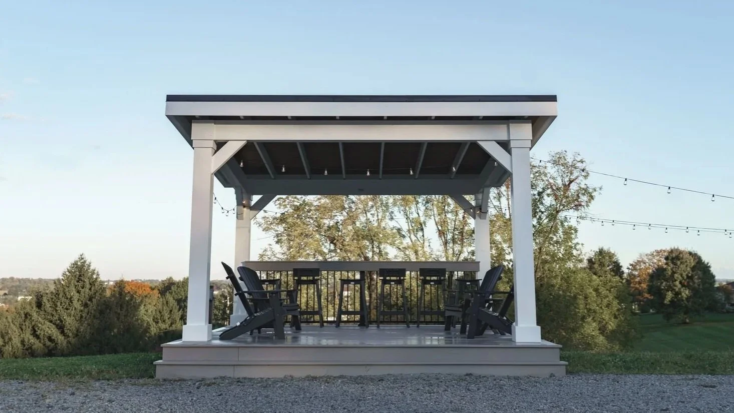 Outdoor pavilion with a bar counter and six chairs, set in a grassy area with trees and string lights.