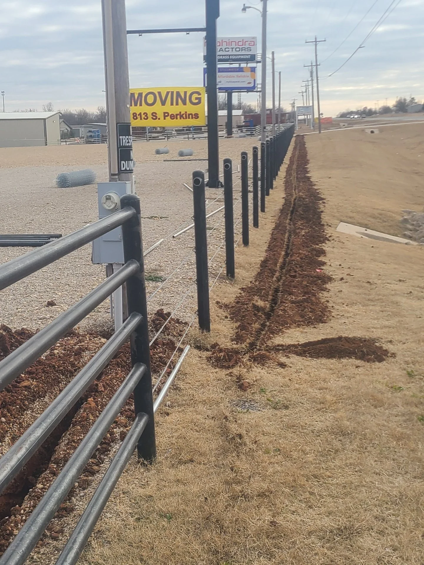 Construction site with a black metal fence, a moving sign with an orange and yellow background, and a row of utility poles along a dirt pathway.