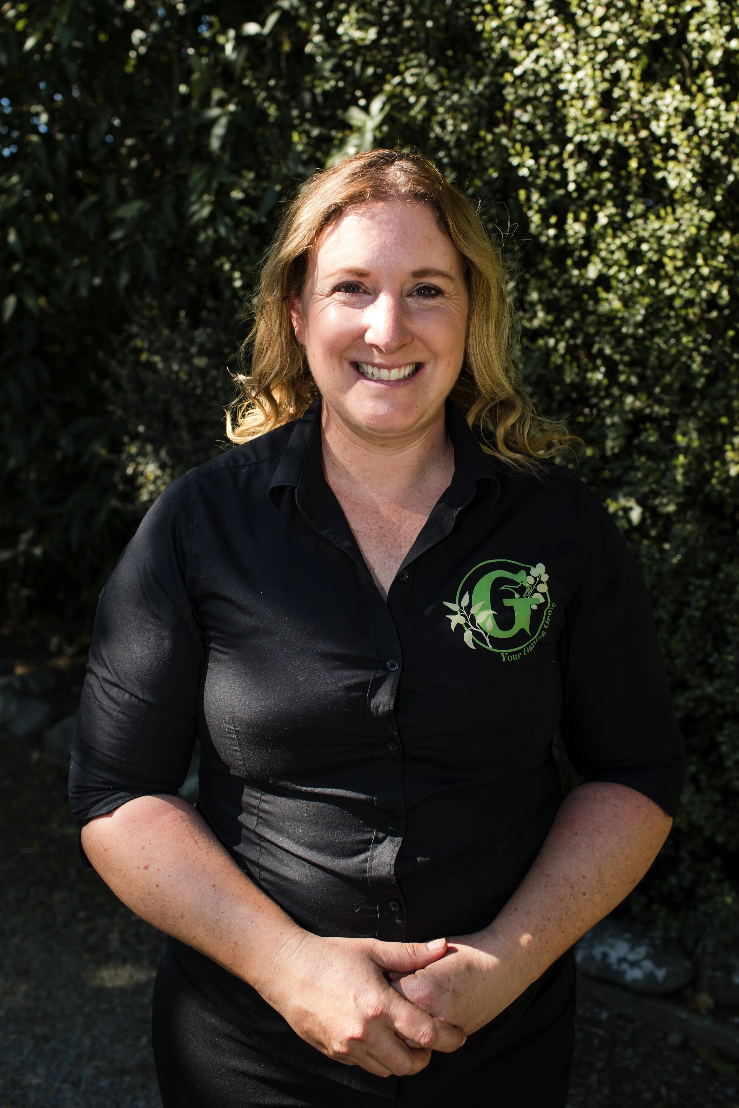 A smiling woman with shoulder-length blonde hair outdoors, wearing a black button-up shirt with a green logo on the left chest.