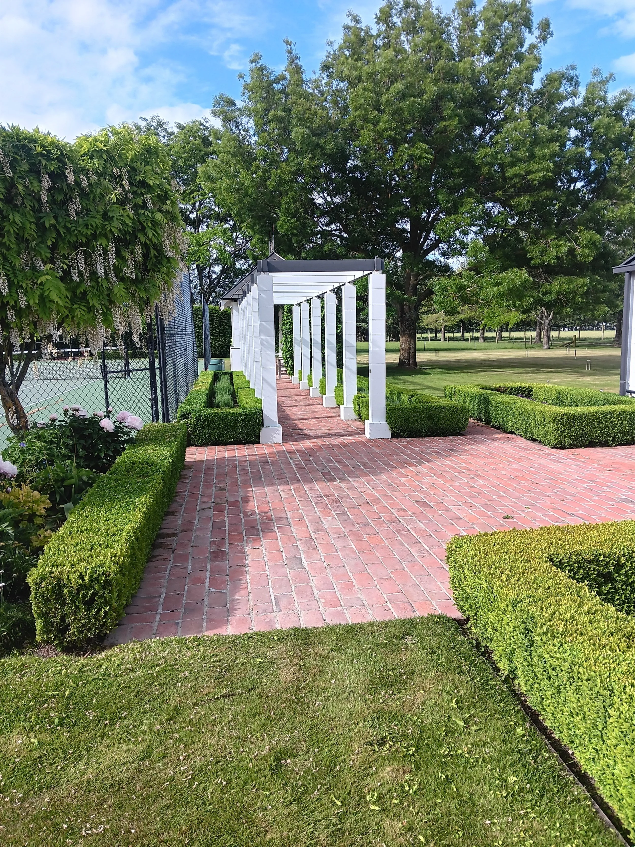 Garden with a brick walkway and white pergola, surrounded by trimmed bushes and trees, near a fence and tennis court.