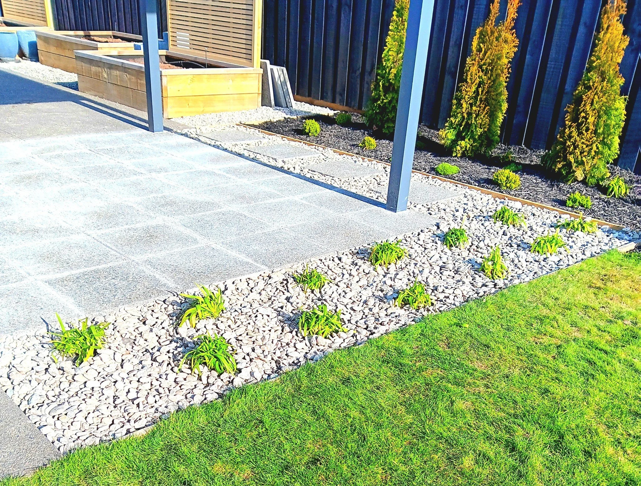 A neatly landscaped backyard garden with a patio, stone walkway, and green grass, featuring small shrubs and trees along a dark wooden fence.