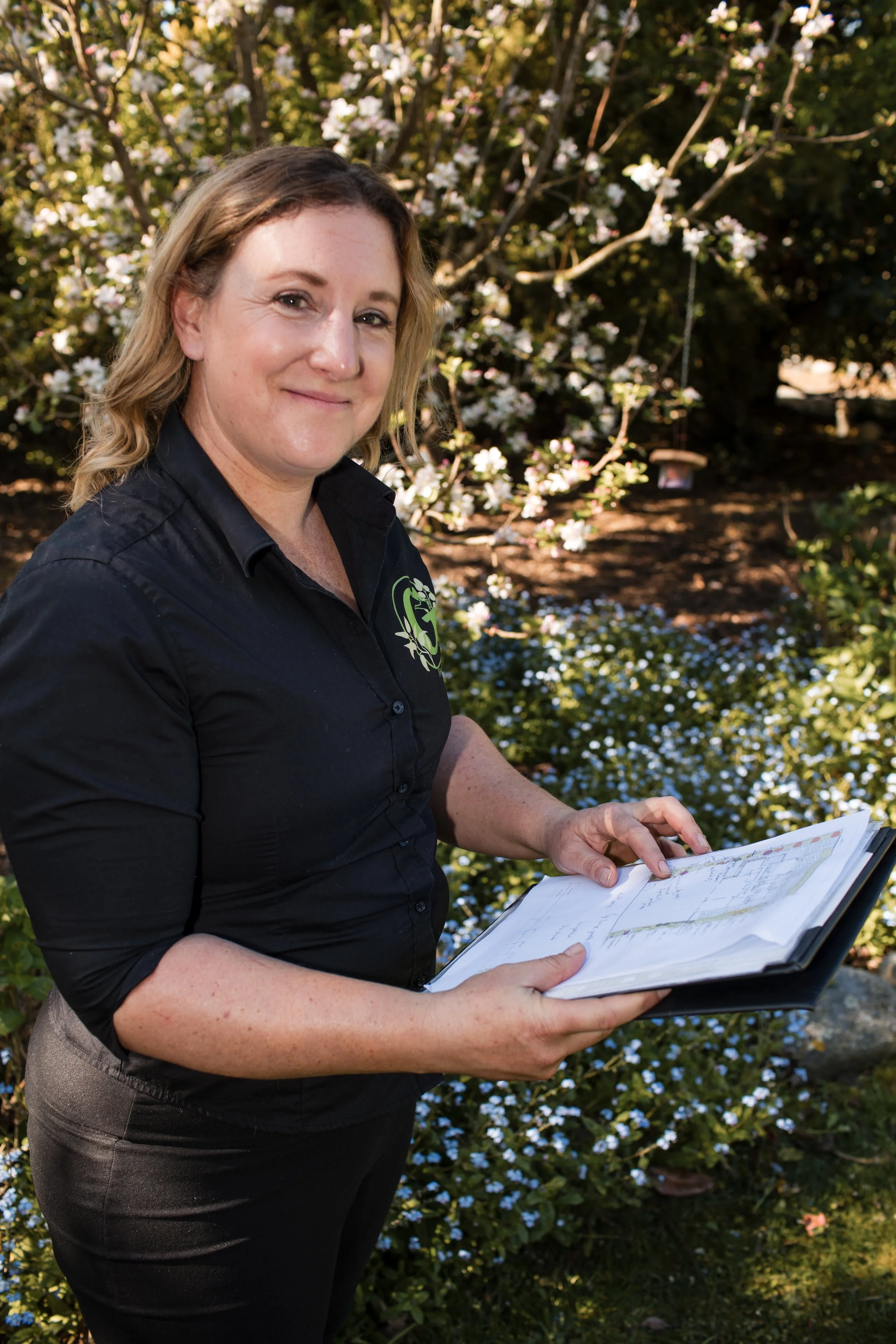 A woman in a black shirt with a green logo holding an open book outdoors, with flowering bushes and small white flowers in the background.
