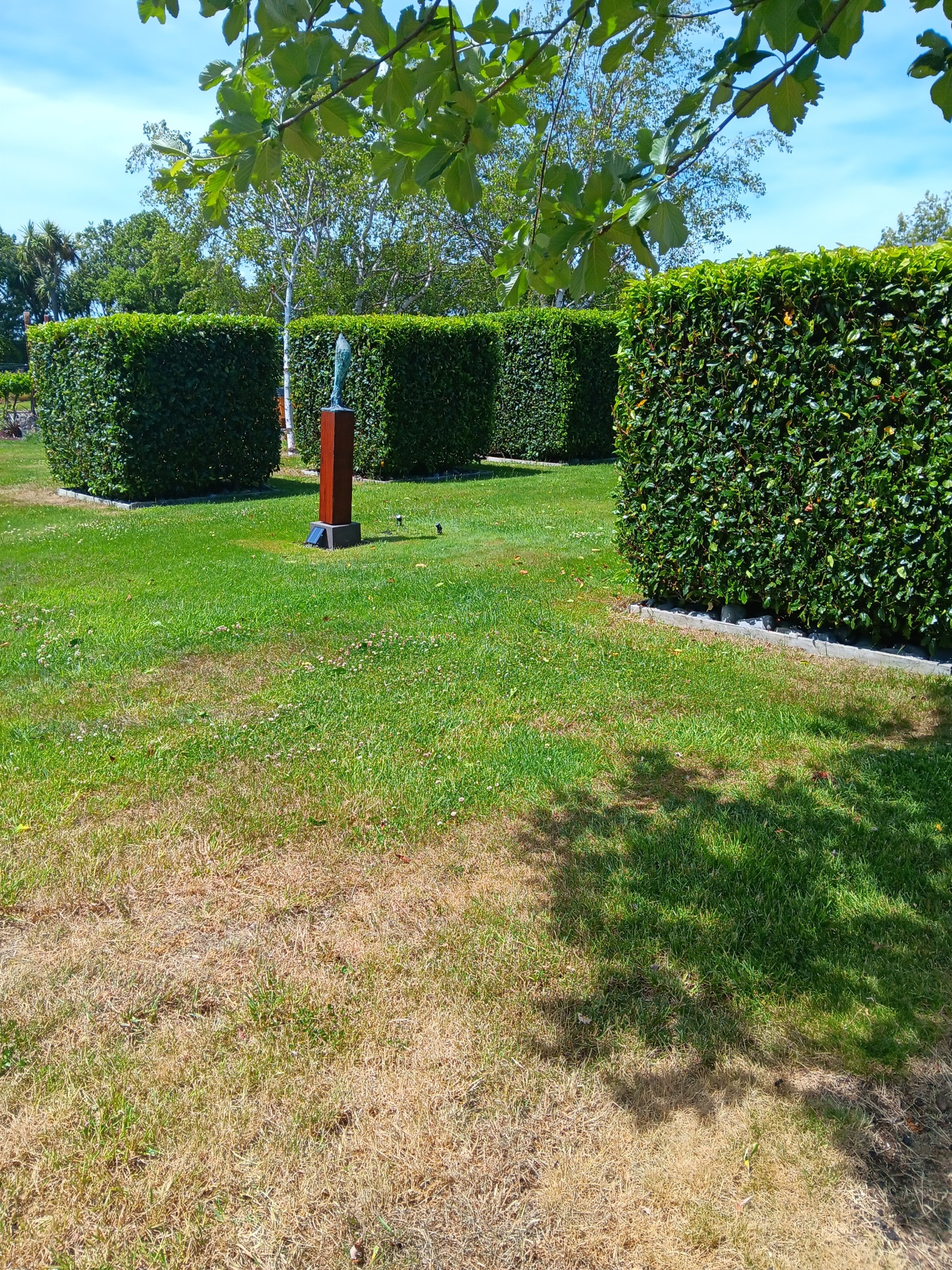 A well-maintained grassy park with neatly trimmed hedges and painted sculptures. Trees and a clear blue sky are visible in the background.