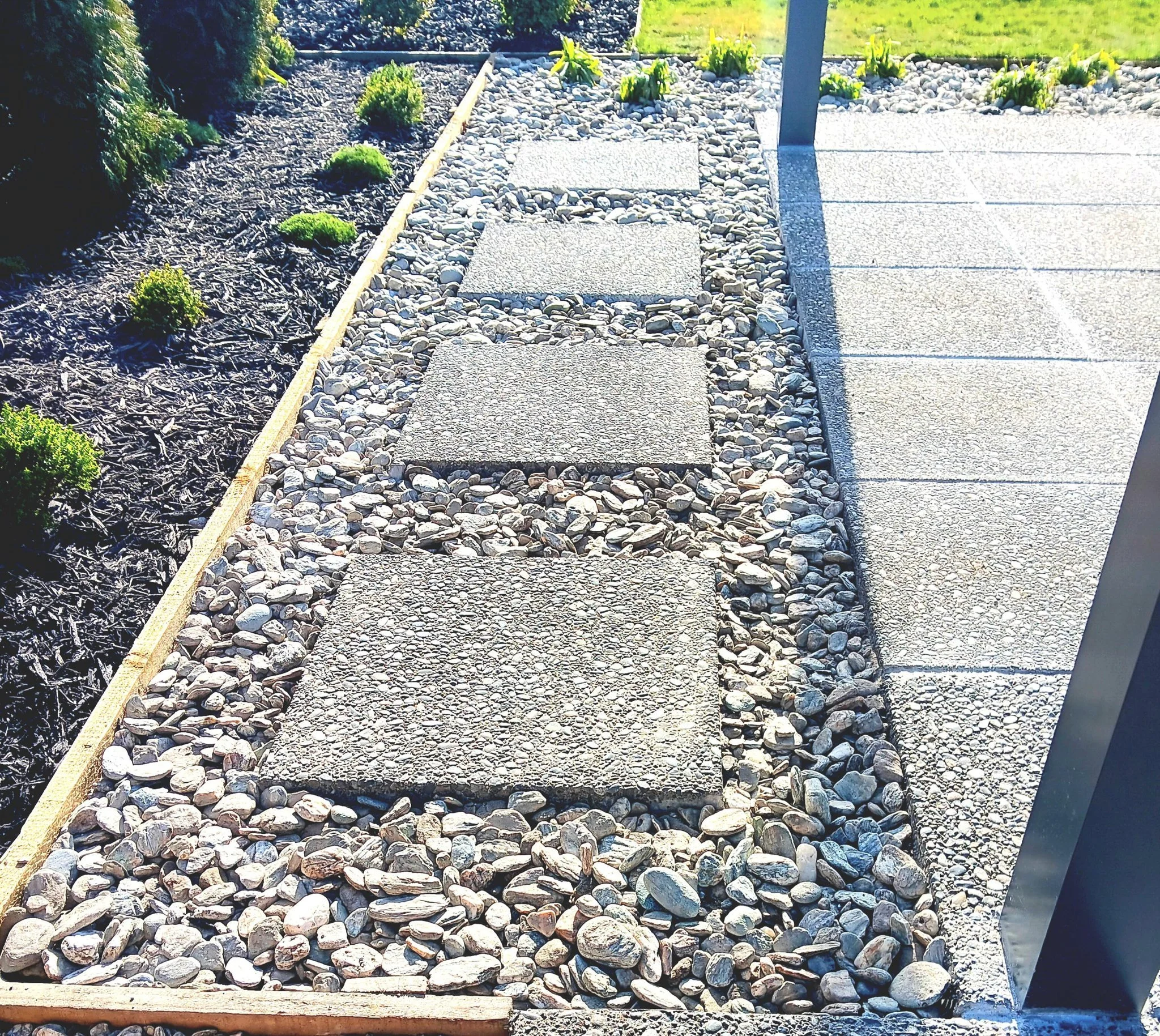 Garden pathway with square concrete stepping stones and small decorative rocks, bordered by a wooden edging, next to a landscaped garden bed with small green shrubs and mulch.