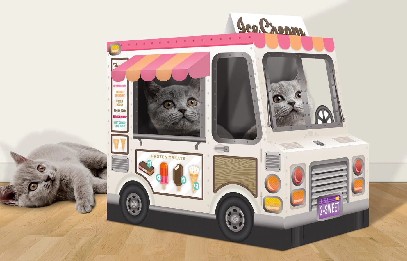 A grey kitten lying on a wooden floor next to a toy ice cream truck with two other grey kittens inside, looking through the windows.