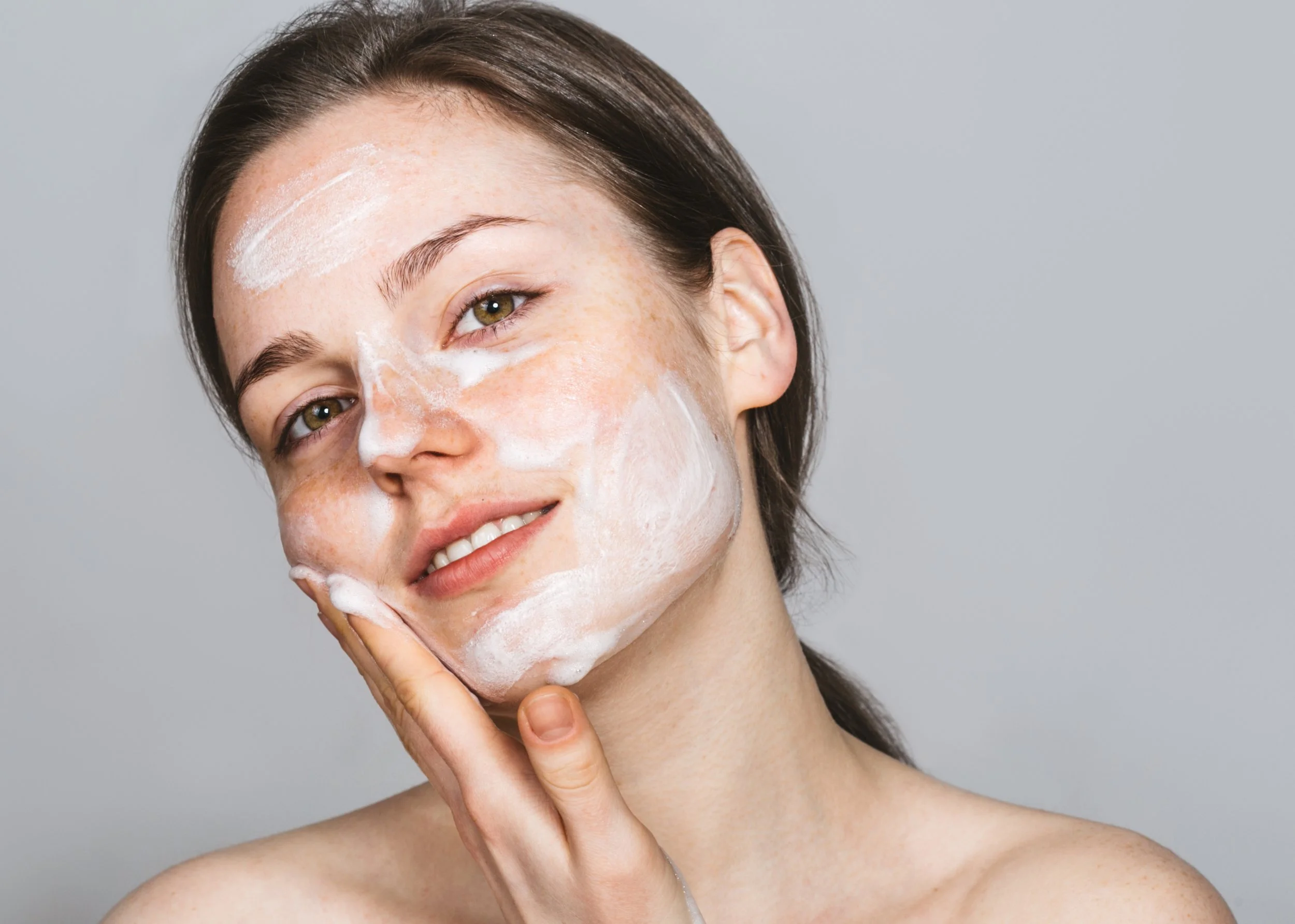 Woman with short brown hair applying a foamy facial cleanser or mask to her face, touching her cheek with her hand, against a neutral background.