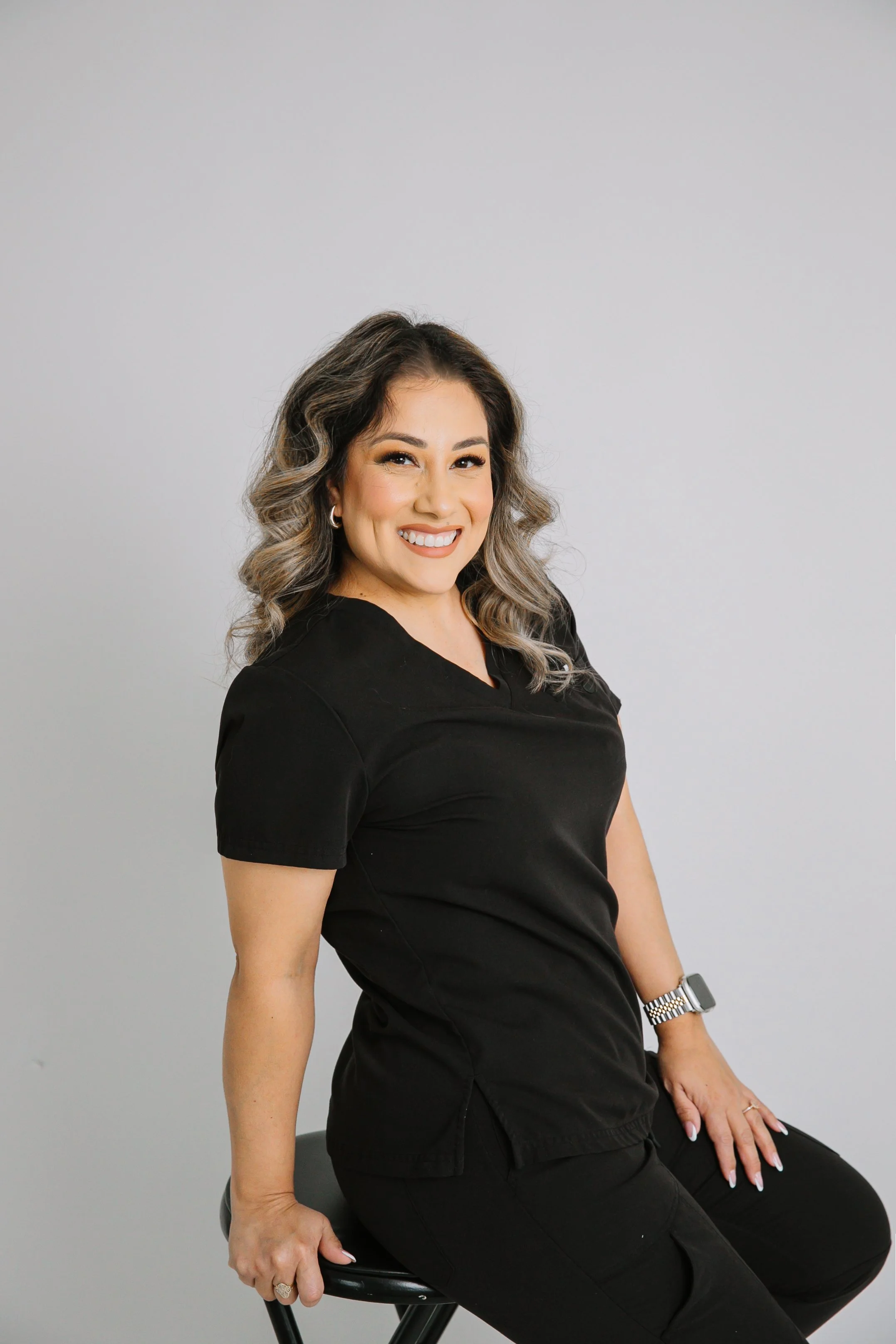 Licensed esthetician seated on stool, smiling in professional black scrubs against a neutral studio background