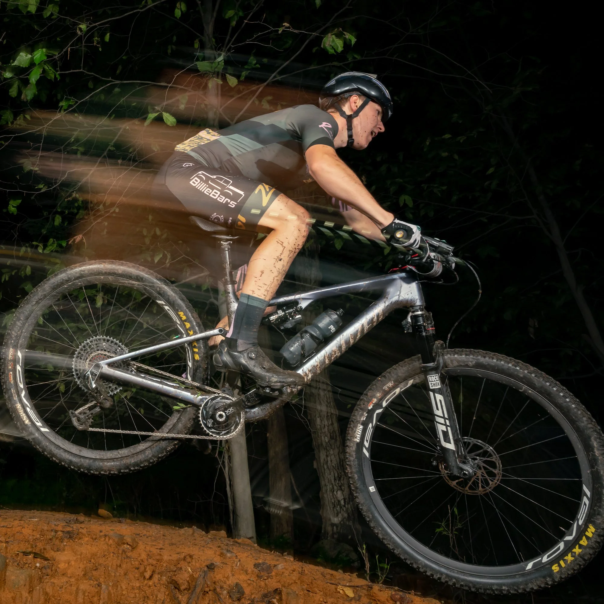 A mountain biker in motion, riding uphill on a trail at night, with a dark background and some trees, wearing a helmet and athletic gear.