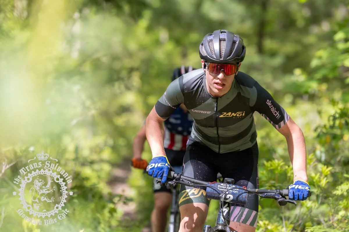 Cyclist in black and gray racing gear, wearing a black helmet and red sunglasses, biking through a green forest trail.
