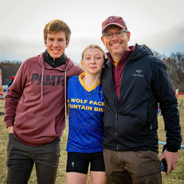 Three people standing outdoors, smiling at the camera. The person in the middle is a young woman wearing a blue sports jersey with yellow lettering, the person on the left is a young man in a maroon hoodie, and the person on the right is an older man wearing glasses, a maroon cap, and a black jacket.