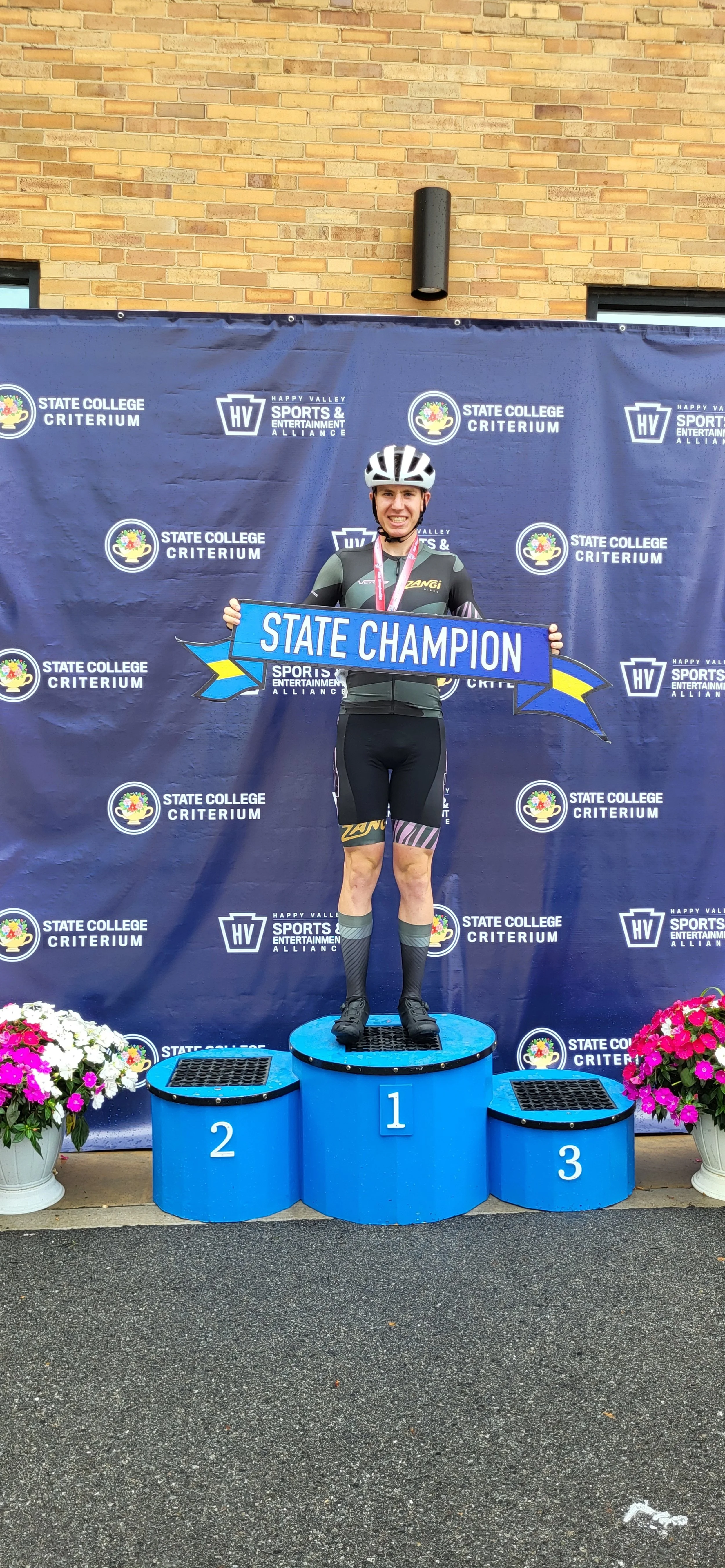 A cyclist standing on the first-place podium holding a banner that reads 'State Champion,' smiling, with a medal around their neck, in front of a backdrop with event logos, and two potted flowers on either side.