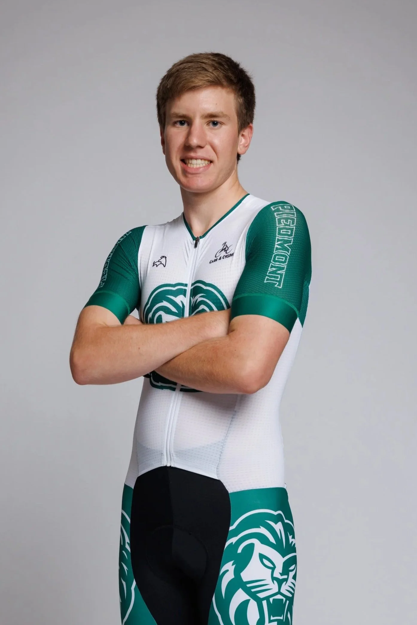 A young male cyclist posing with arms crossed, wearing a white and green cycling jersey and black cycling shorts with a lion logo, against a plain gray background.