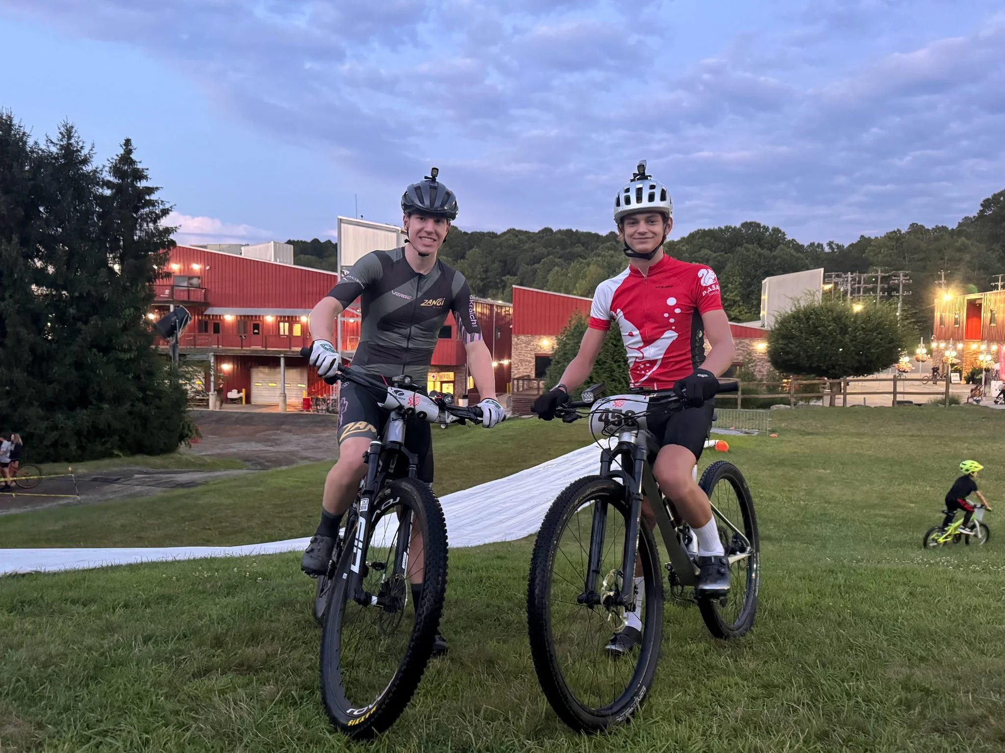 Two young men in cycling gear and helmets posing with mountain bikes on a grassy area at dusk, with a red building and other cyclists in the background.