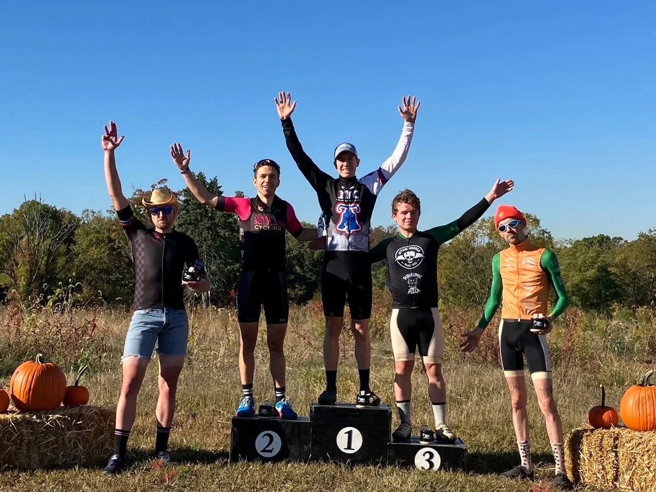 Five cyclists in racing gear standing on a winners' podium outdoors, with pumpkins and hay bales around, celebrating a race with a blue sky and trees in the background.