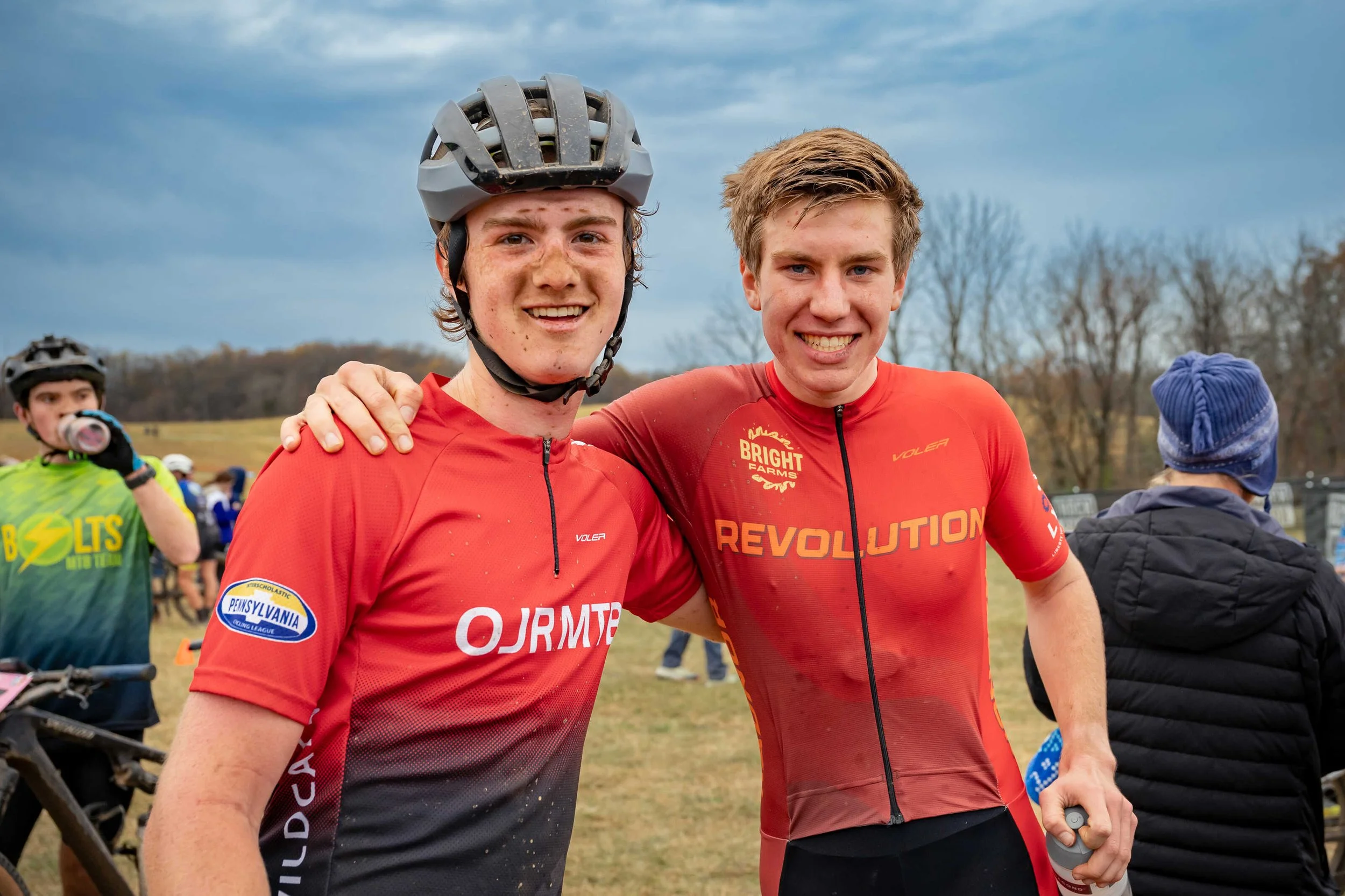 Two young men in red cycling jerseys smiling and posing for a photo outdoors, with other cyclists and trees in the background.