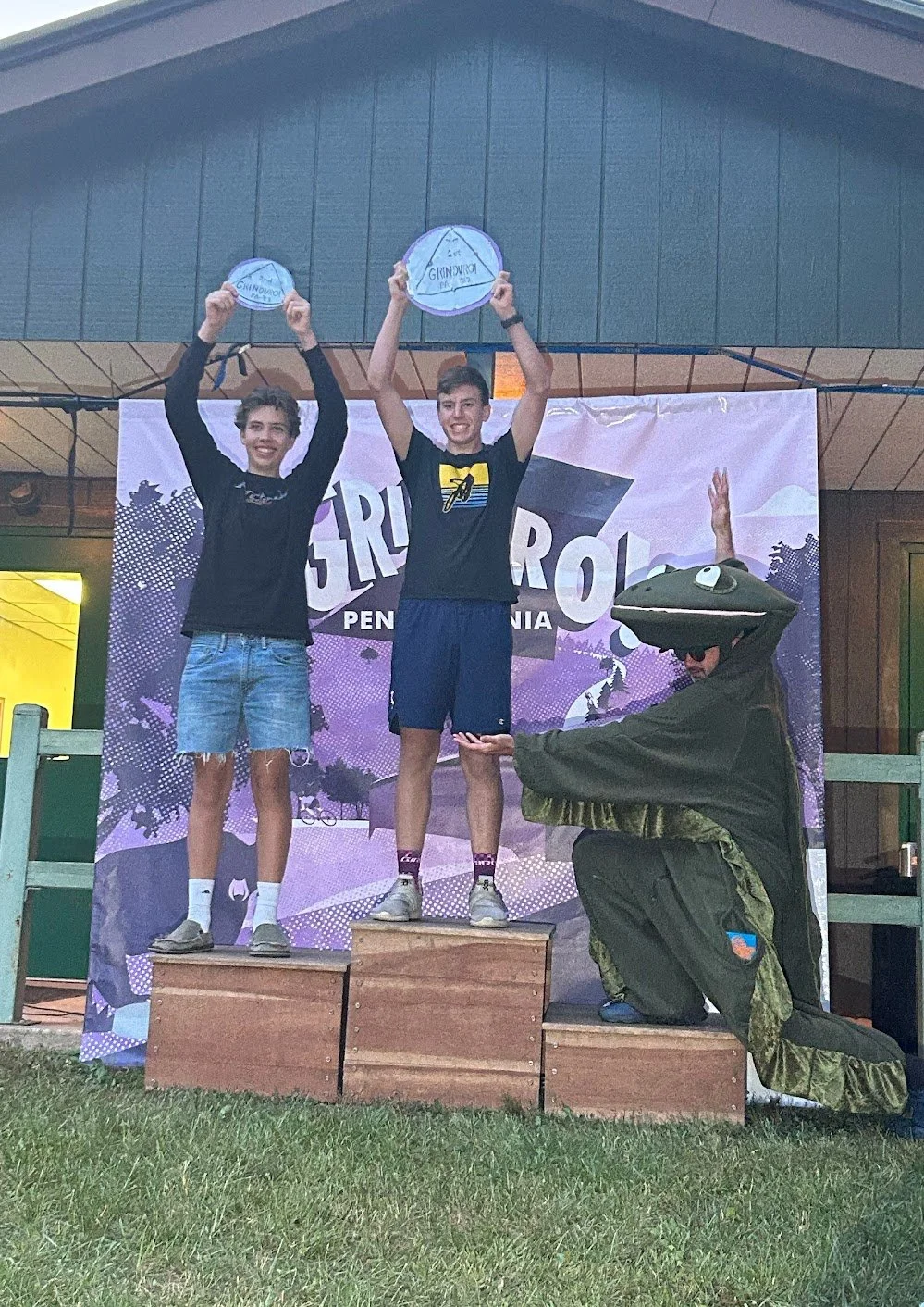 Two boys standing on top of a winners' podium, holding awards above their heads, while a person in a crocodile costume kneels beside them, pointing at their trophies. The background features a purple event banner.