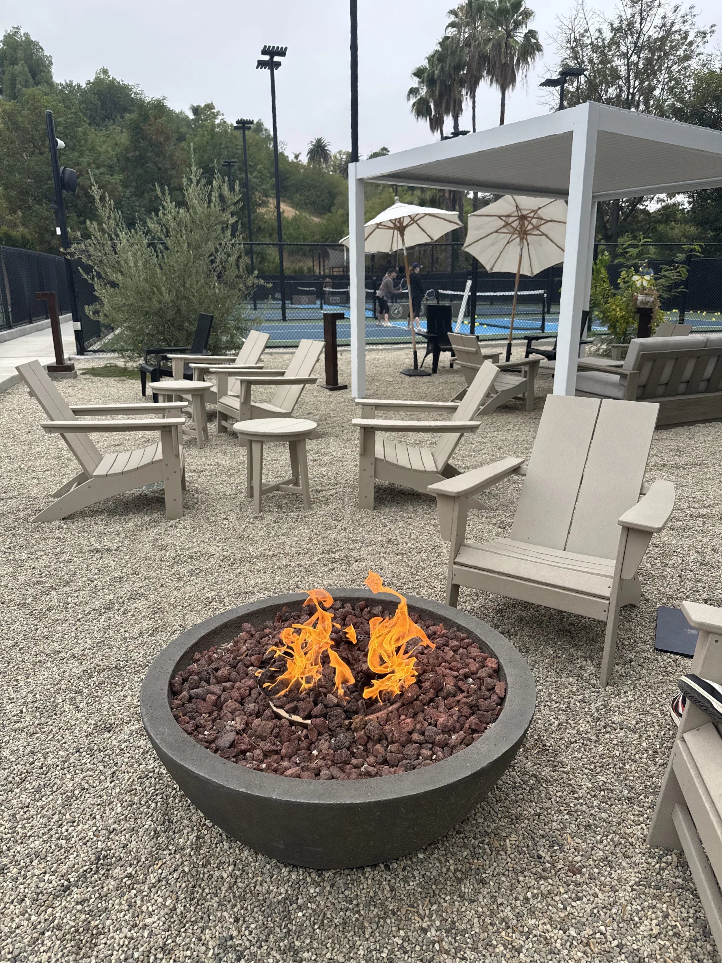 Outdoor lounge area with beige chairs, small tables, a fire pit, a tennis court in the background, and patio umbrellas under an overcast sky.