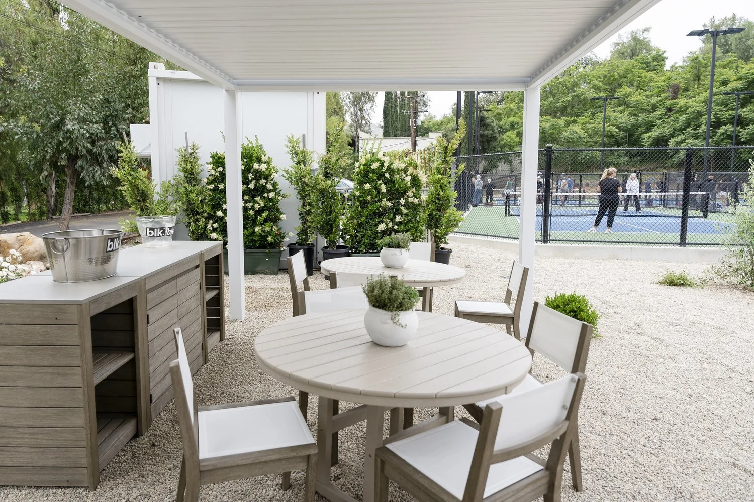 Outdoor patio with white round tables, beige chairs, potted plants, and a tennis court in the background with people playing.