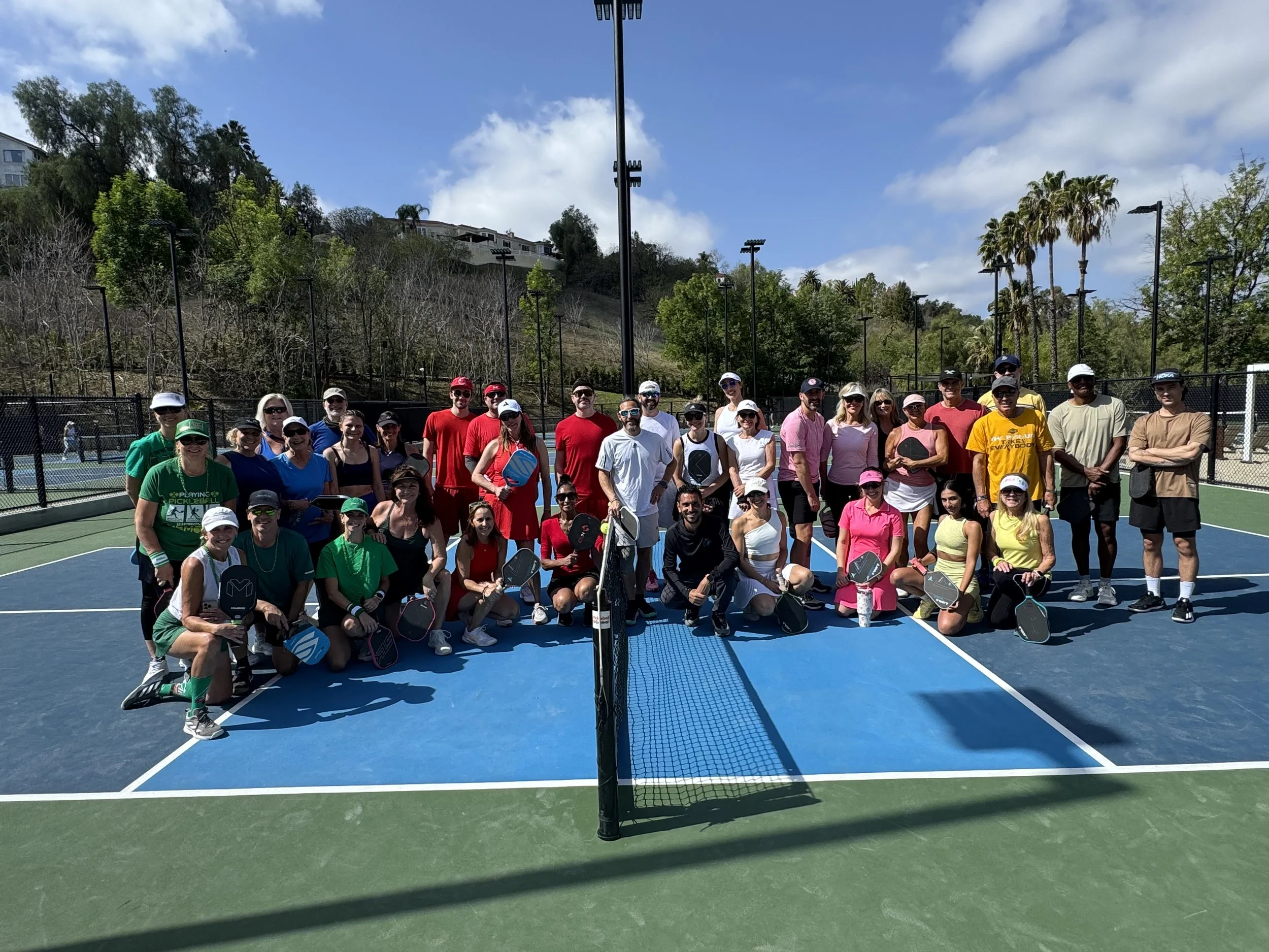 A large group of people on a tennis court, holding paddles, posing for a group photo under a sunny sky with some clouds, surrounded by trees and a hillside.