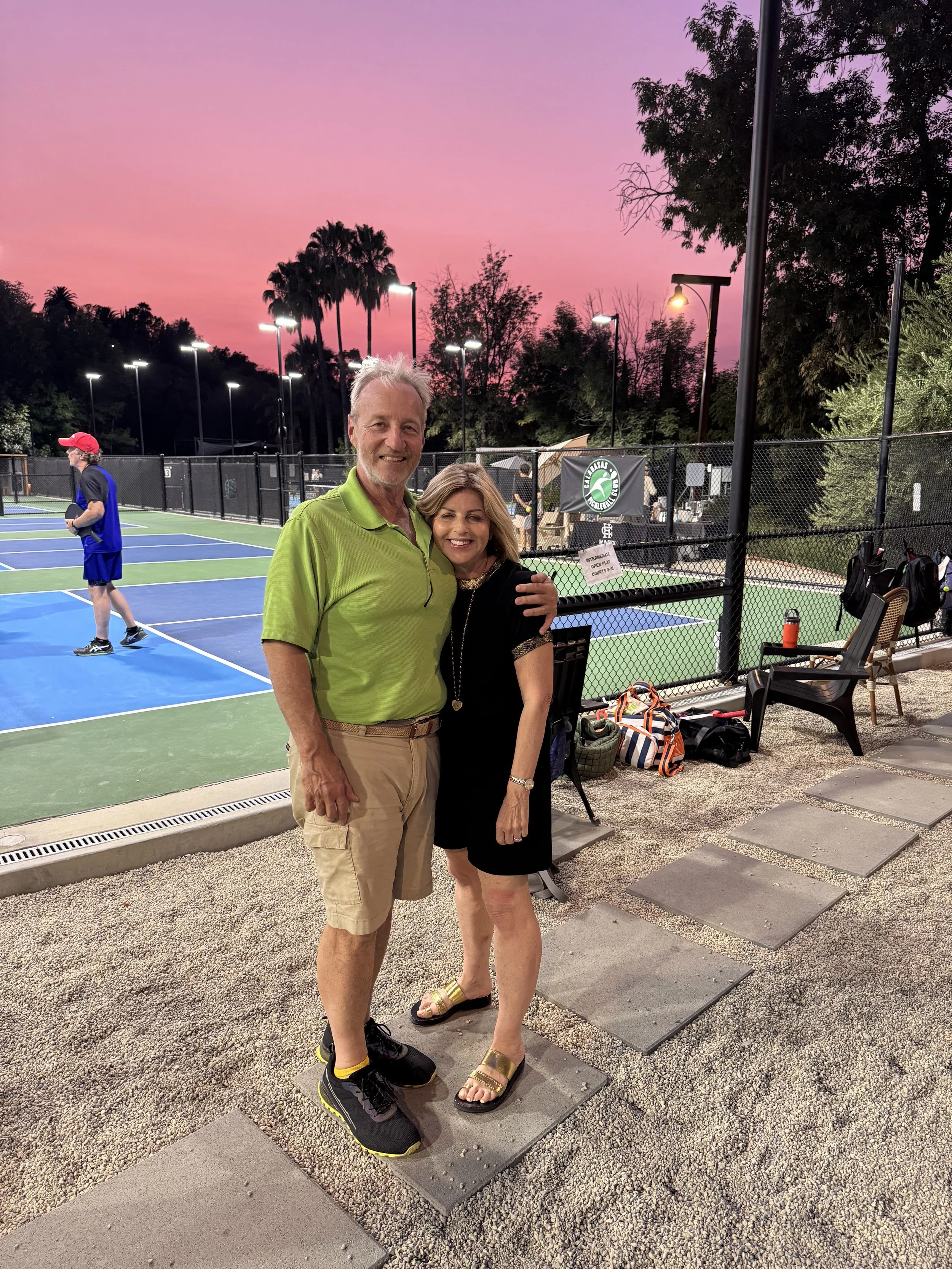 Two people, a man and a woman, smiling and hugging at a tennis court during sunset with pink sky, blue and green tennis courts, and trees in the background.