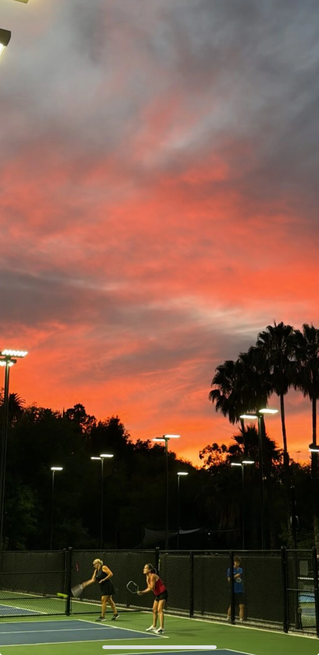 Sky at sunset with pink and orange clouds over a tennis court where three women are playing, with palm trees and tall lights in the background.