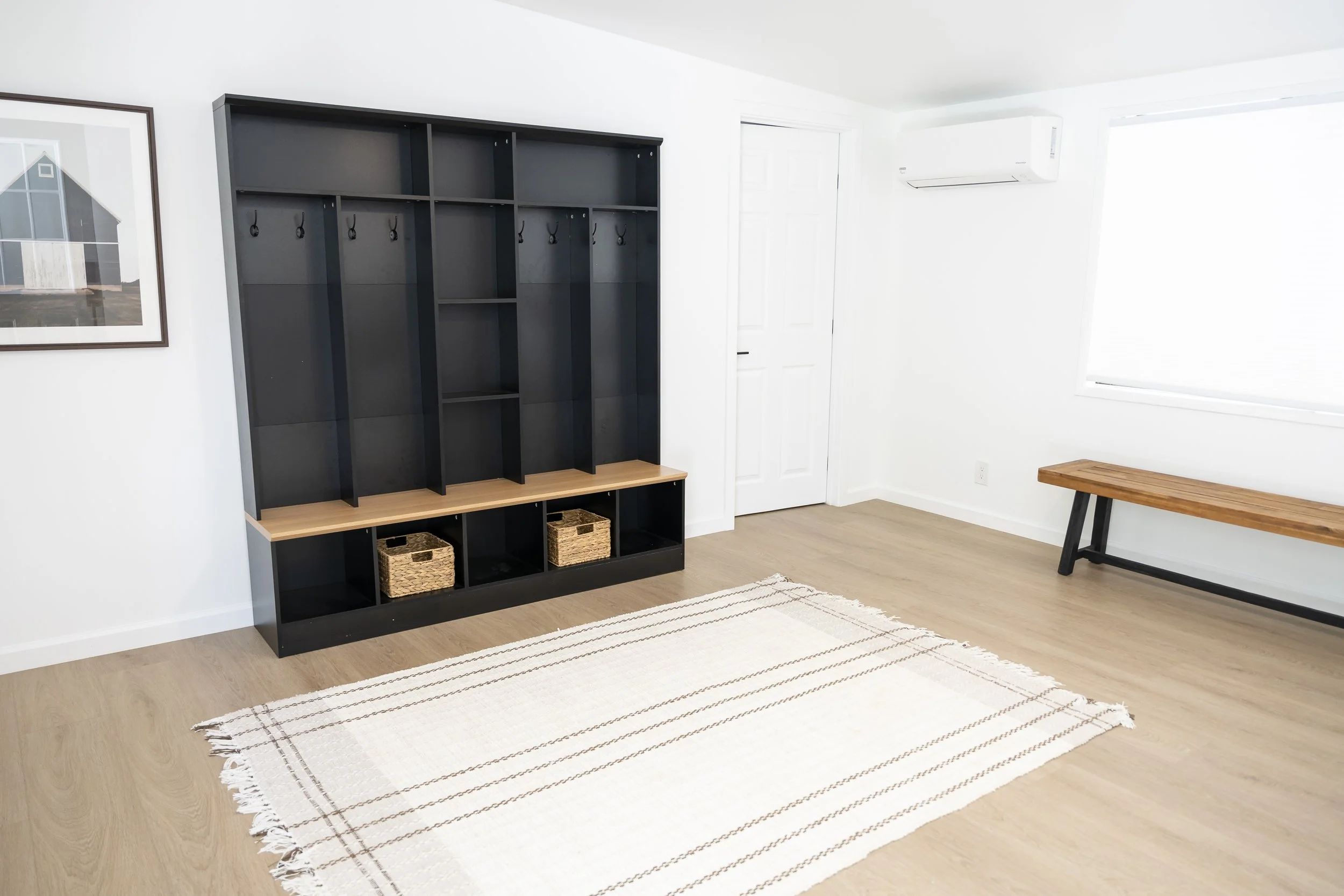 Empty living room with black and wood coat rack, wooden bench, white walls, light wood flooring, window, air conditioning unit, framed artwork, white door, and striped rug.