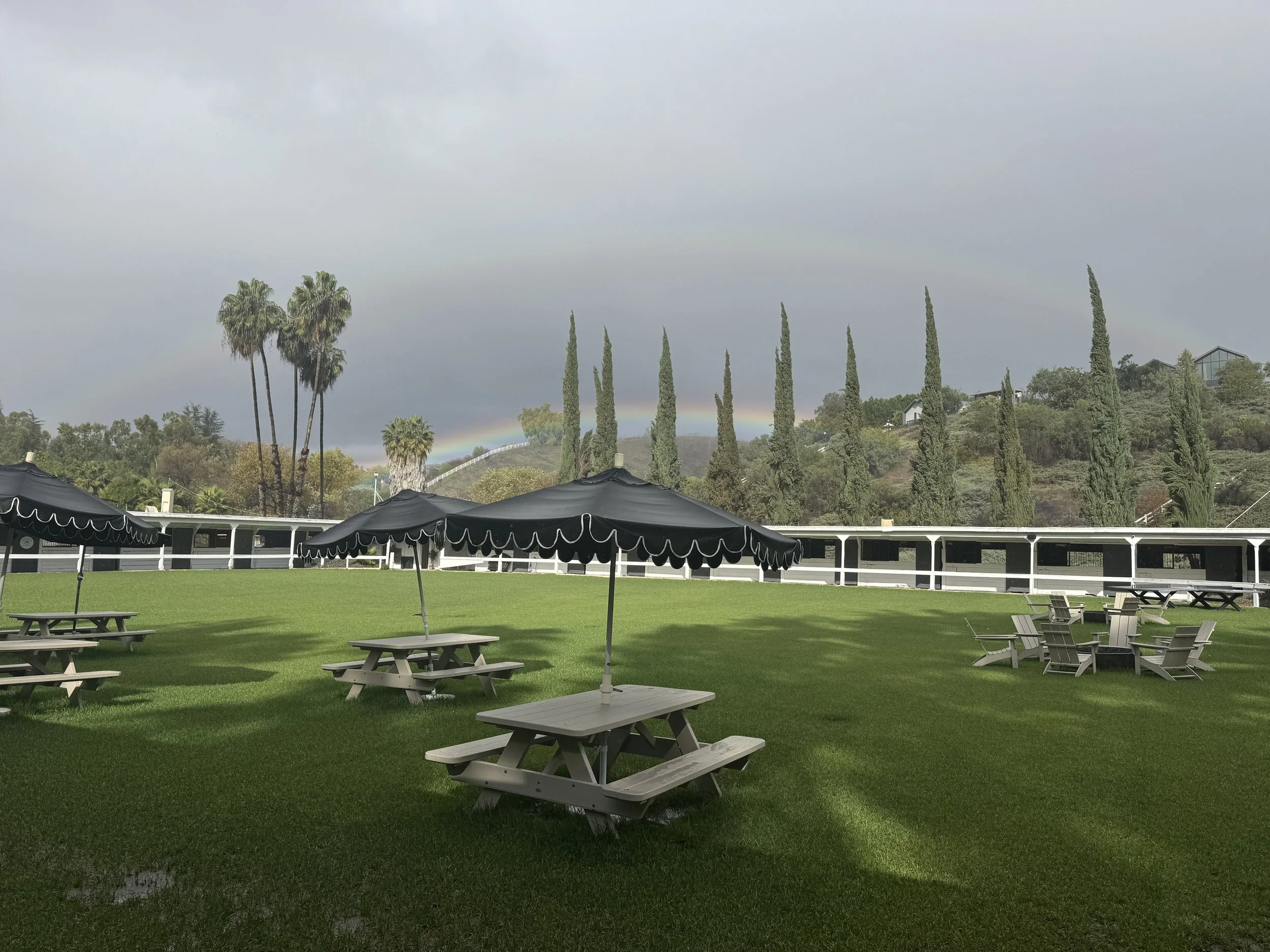 An outdoor grassy area with picnic tables and umbrellas, trees, hills, and a rainbow in the background.