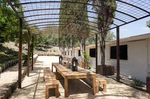Outdoor picnic area with a wooden table and benches under a curved metal canopy, surrounded by trees and a white building.