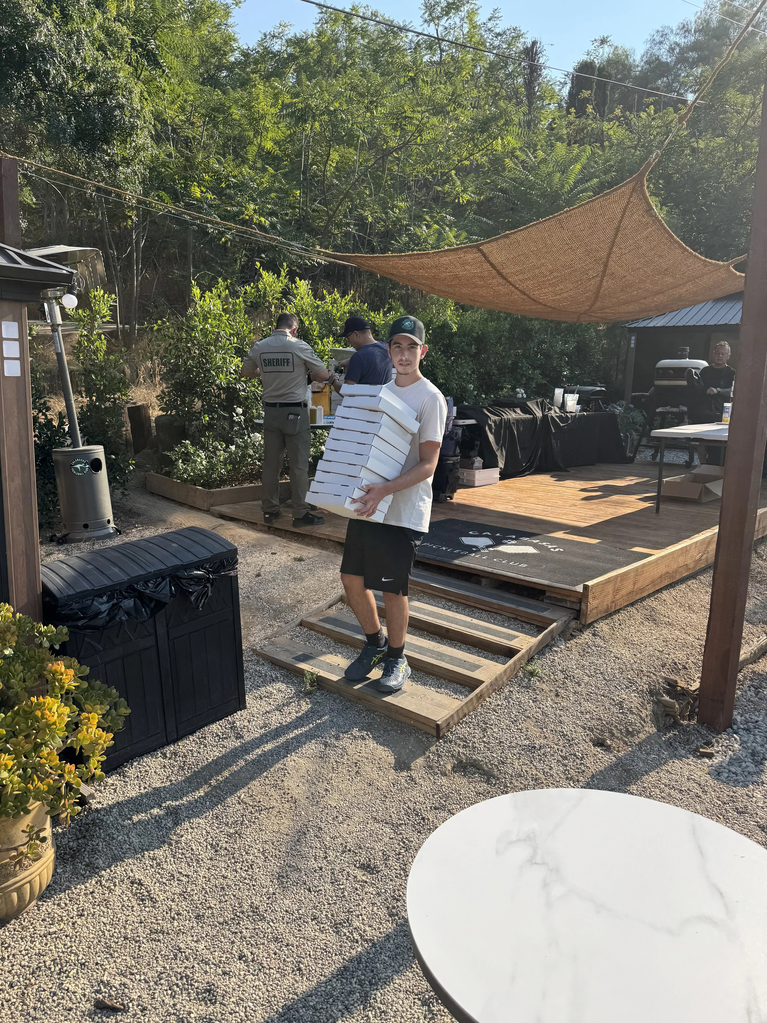 A young man wearing a white t-shirt, black shorts, and a black cap holding a stack of pizza boxes outdoors. In the background, several people, including a sheriff, are preparing food at a table under a shade sail. There is a trash bin and a white cir