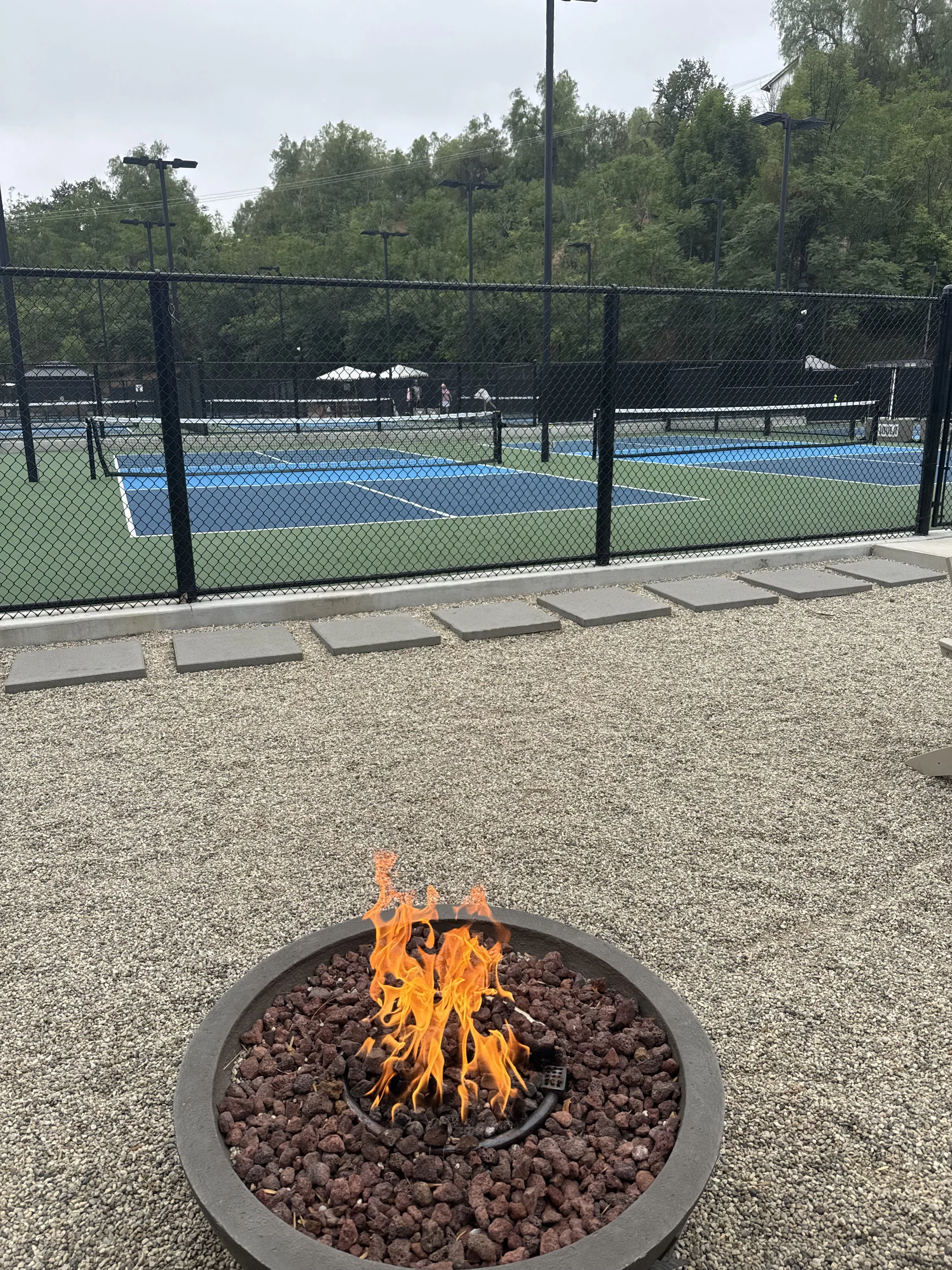 A fire pit with a small flame in the foreground, a gravel surface around it, and a fenced outdoor tennis court with blue and green surfaces in the background. There are trees beyond the netted fencing.