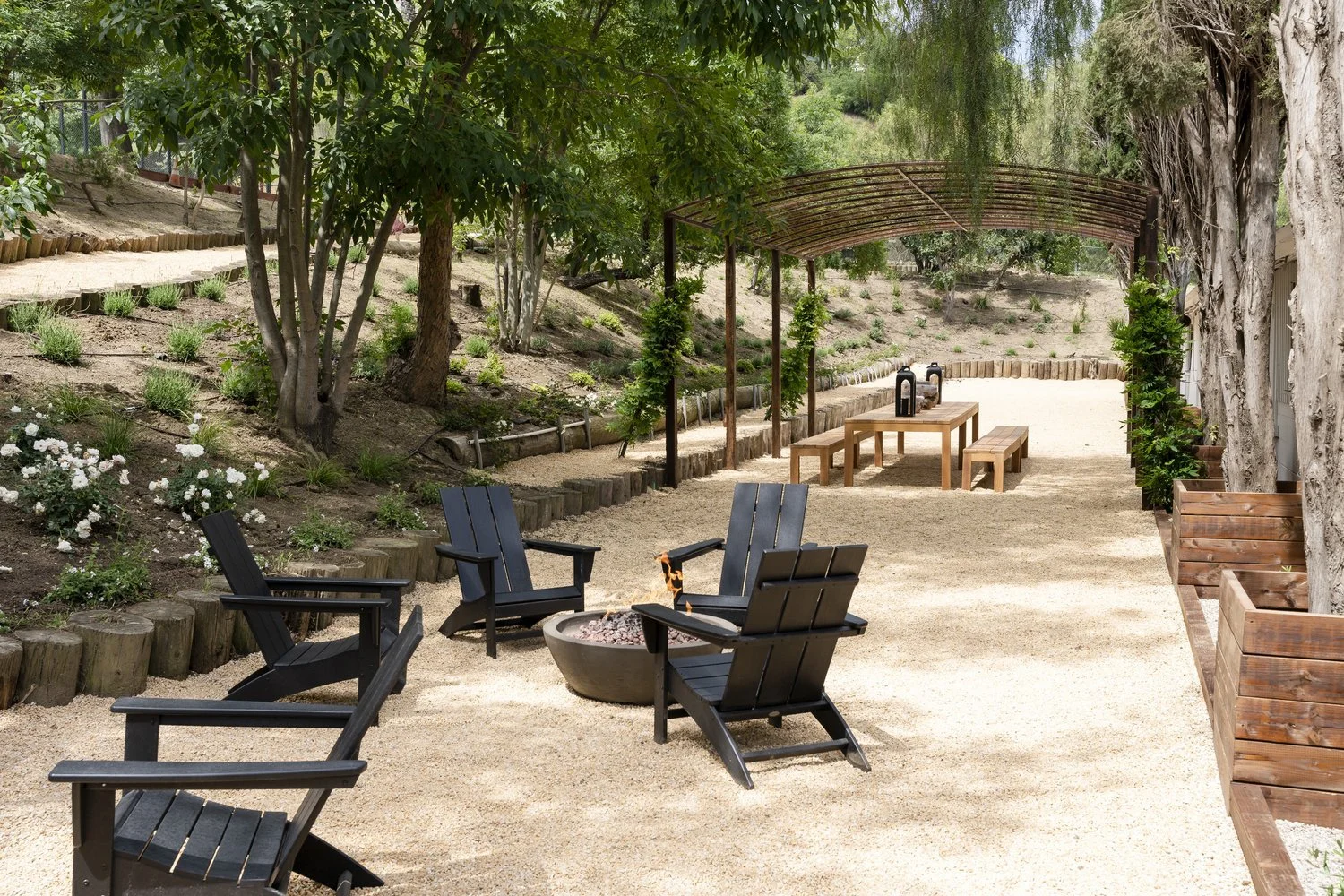 Outdoor seating area with black Adirondack chairs surrounding a fire pit, wooden picnic table underneath a metal arbor, and a shaded area with two bottles on the table. Landscaped garden bed with white flowers and trees on the left, and a gravel path
