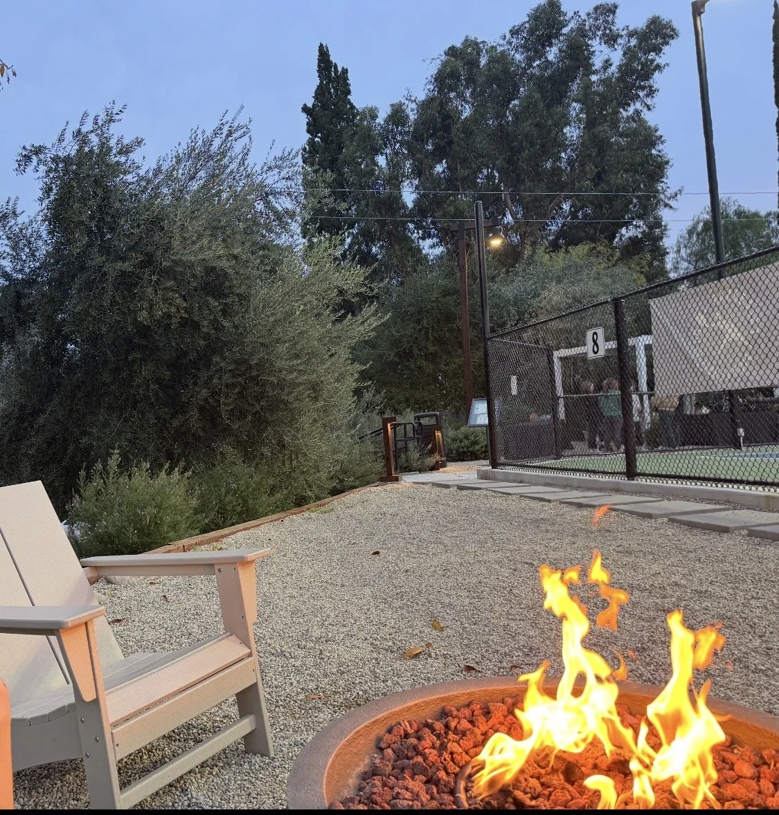 An outdoor scene showing a fire pit with a small flame, a bench to the left, and a fenced tennis or pickleball court in the background. Tall trees and bushes surround the area, and a few lights are visible on poles near the court.