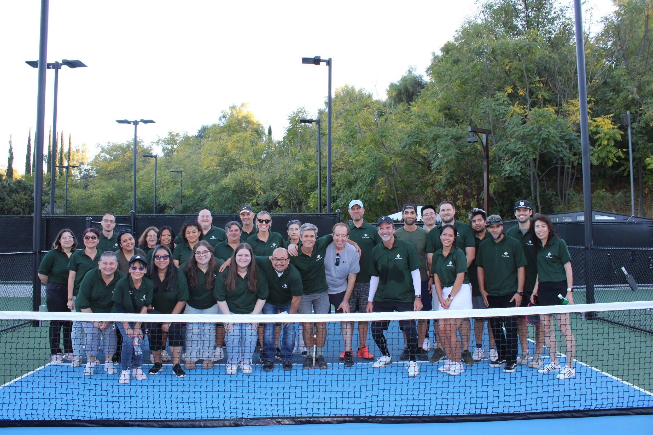 Group of people standing on a tennis court, some smiling at the camera, wearing matching green polo shirts, with trees and outdoor lighting in the background.