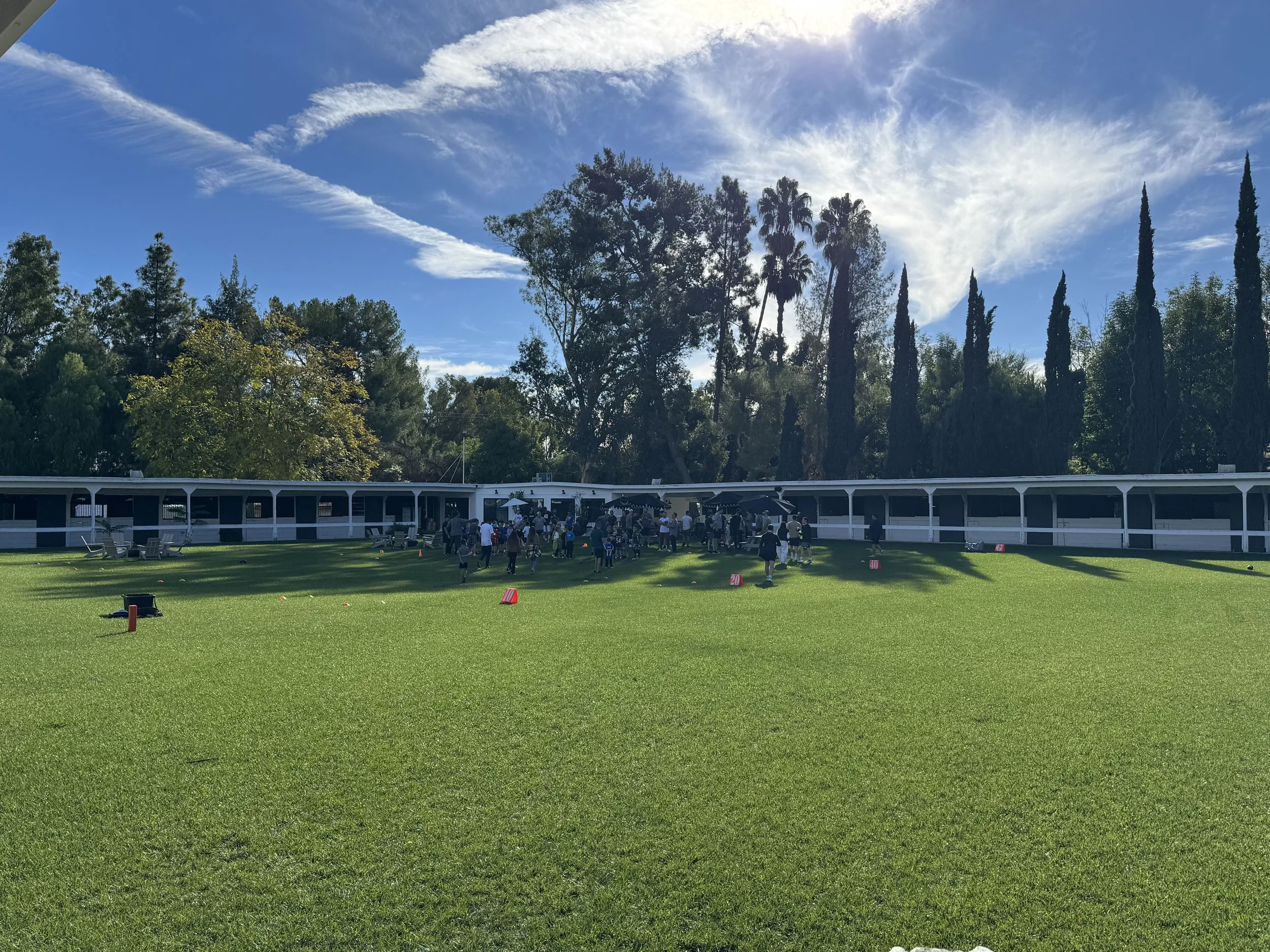 A group of people gathered on a football field with a white building and tall trees in the background under a blue sky with clouds.