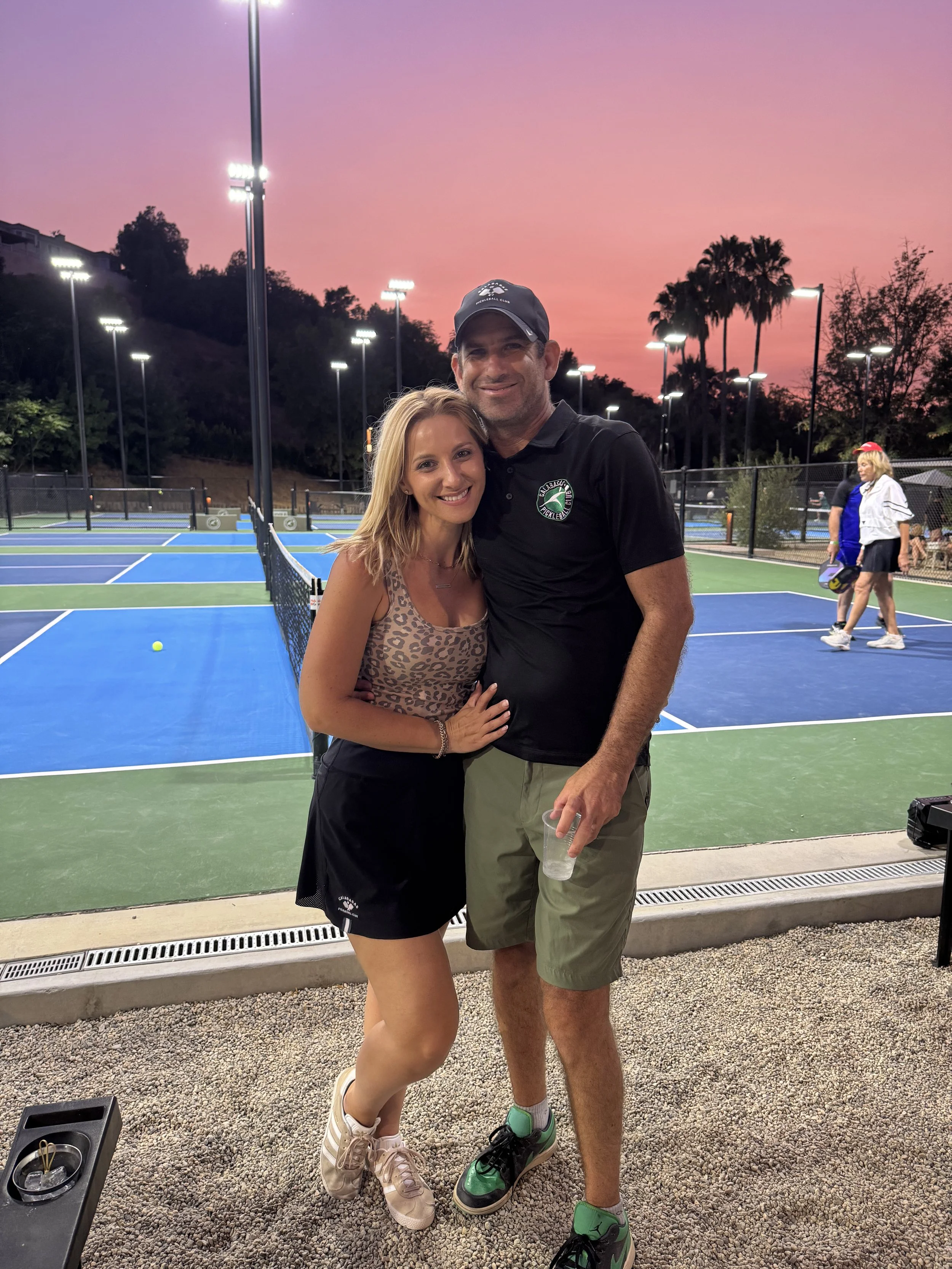 A smiling woman in a leopard print tank top and black shorts stands next to a smiling man in a black polo shirt and khaki shorts, holding a plastic cup, at an outdoor tennis court during sunset.