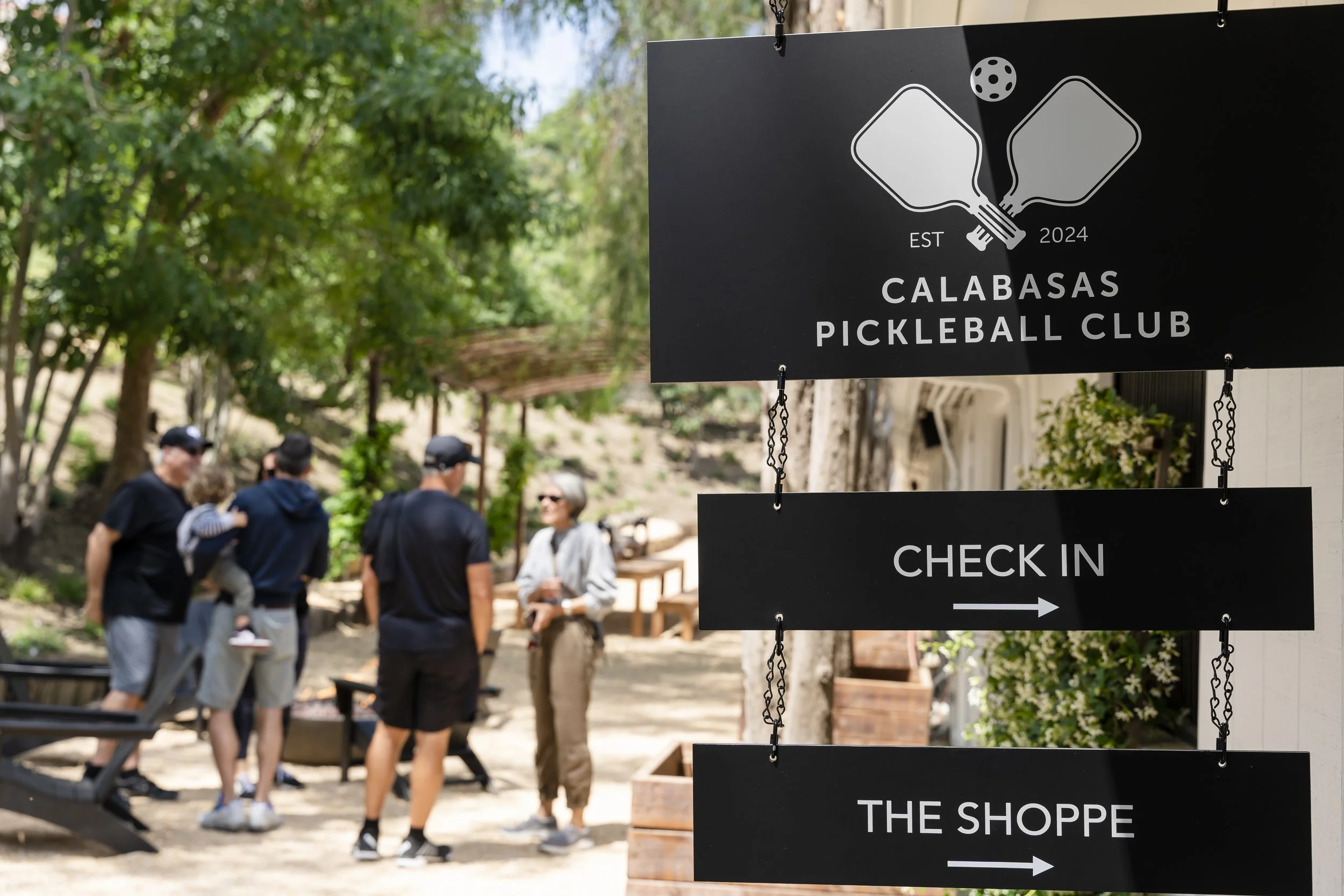 Sign for Calabasas Pickleball Club with check-in instructions, referencing a pickleball paddle and ball, in a park with people standing and talking.