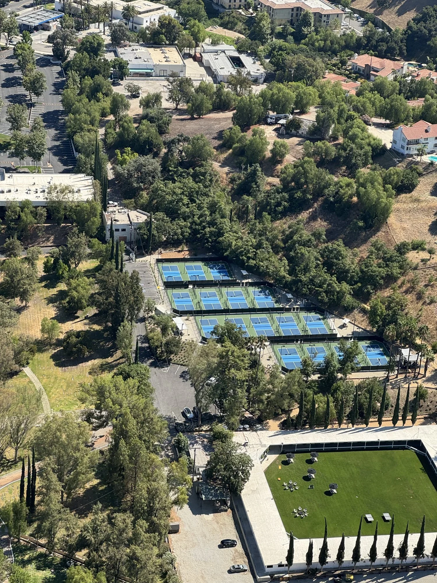 An aerial view of a park with a green lawn, surrounded by trees and a black fence, with outdoor furniture and umbrellas. Adjacent to the park are tennis courts with blue surfaces and a parking lot with some cars. There are residential buildings and g