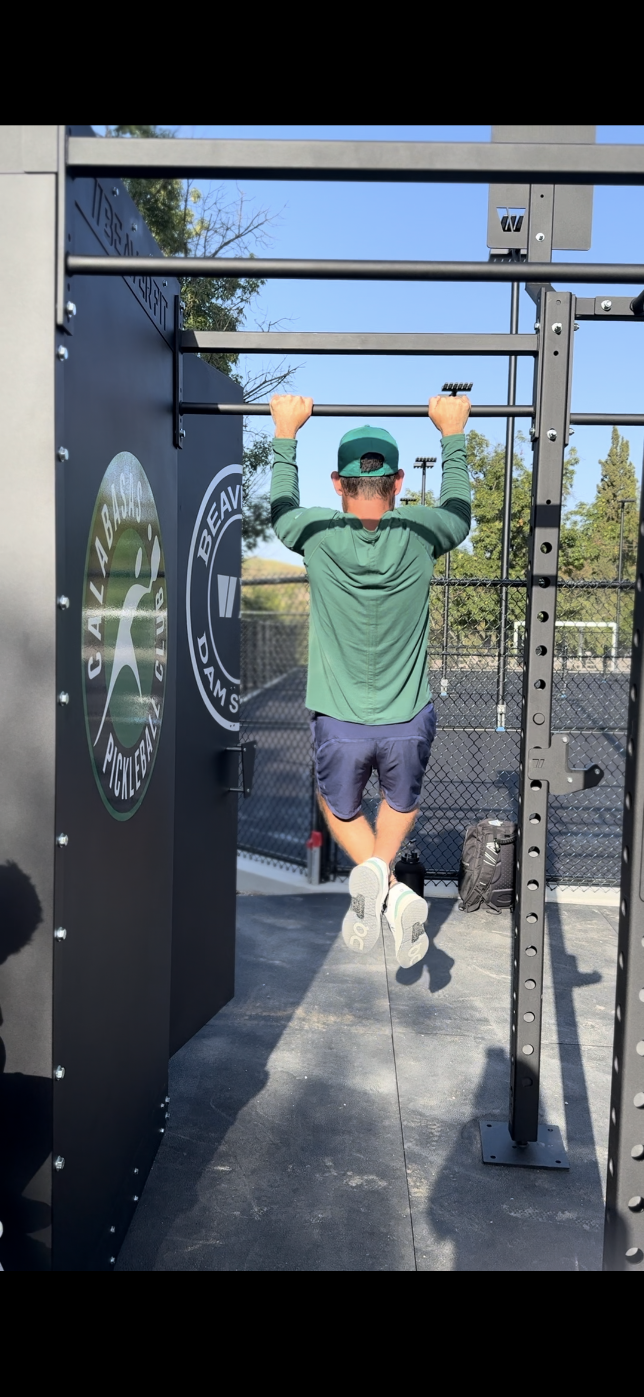 Person wearing a green cap and green jacket doing pull-ups on an outdoor fitness structure.